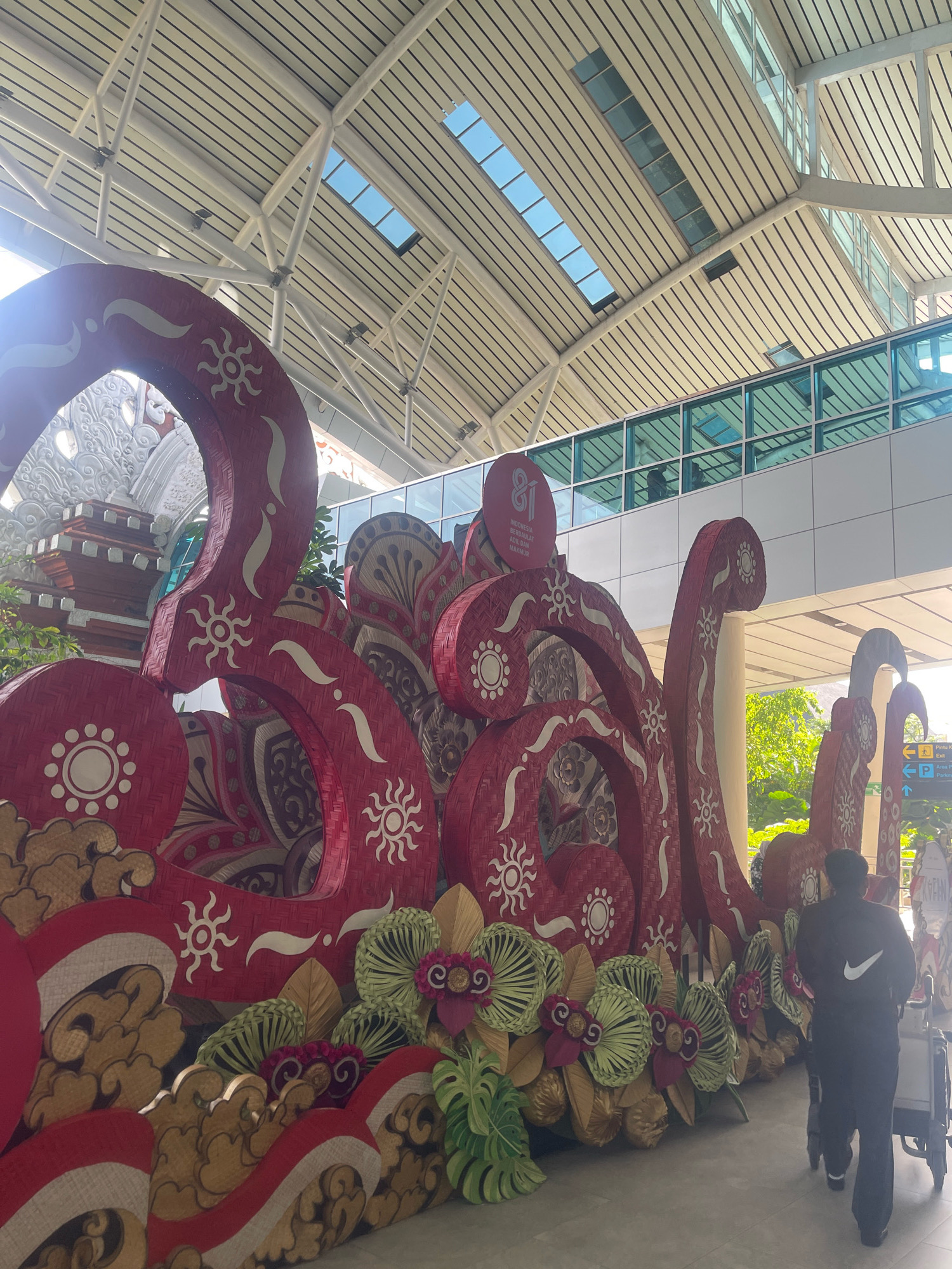 The image depicts a large, ornate, red and white sculpture in what appears to be an airport terminal in Kuta, Indonesia. The sculpture is decorated with intricate patterns, including sun symbols and floral motifs, and is made of woven material. Behind the sculpture, a modern building with large glass windows and a sloped ceiling with skylights is visible. A person is walking away from the camera, pulling a luggage cart, towards the right side of the frame. The lighting suggests it is daytime, with natural light coming through the skylights. The sculpture is likely a decorative installation, possibly for a festival or event, given its elaborate design.