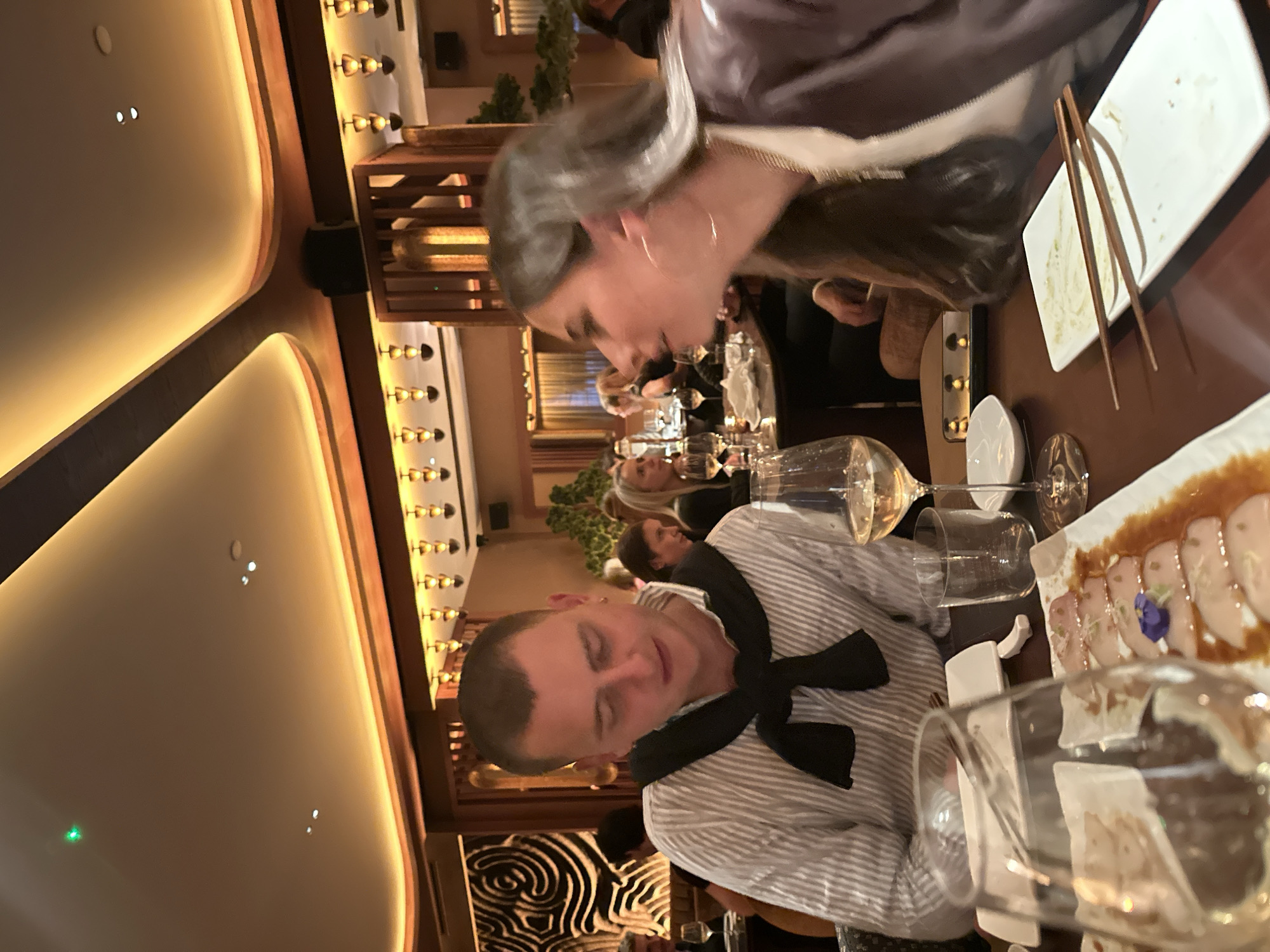 Here is a description of the image:
The image captures a dining scene in a restaurant, likely located in London, United Kingdom, given the composition of the individuals and the ambiance. Two people are prominently featured at the table. One individual, facing the camera, is dressed in a striped shirt with a dark bow tied around their neck. The other, positioned further back, is dressed in a jacket. The table is set with various dishes, including a plate of sliced fish and a plate with chopsticks. Wine glasses are present, suggesting a meal setting. Other individuals can be seen in the background, indicating a bustling restaurant atmosphere. The lighting is warm, creating an intimate setting. Decorative elements such as vertical wooden patterns adorn the surrounding walls and ceiling, further supporting the restaurant environment.