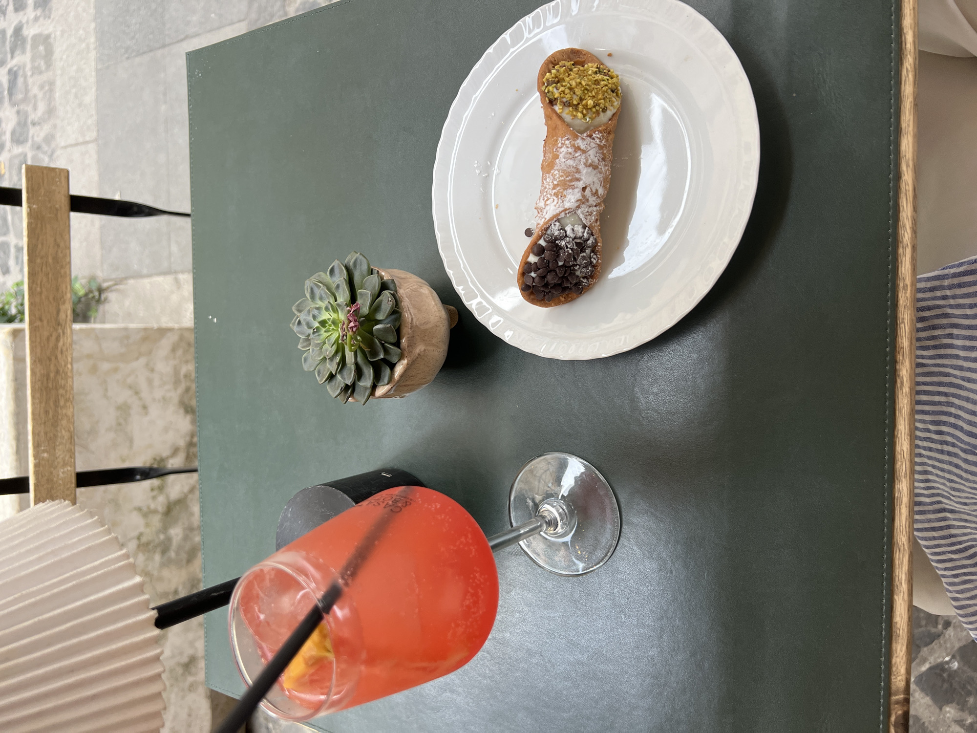 A top-down shot captures a still life on a dark green table. A white plate presents a cannoli, adorned with pistachio at one end and chocolate chips at the other, dusted with powdered sugar. To the left, a succulent in a pot is placed next to a glass of a pink cocktail with a black straw. A striped fabric is visible at the table's edge, and a fragment of a light-colored wicker object is in the lower-left corner. The photo was likely taken at an outdoor cafe in Rome, Italy, given the typical Roman food item "Cannoli".