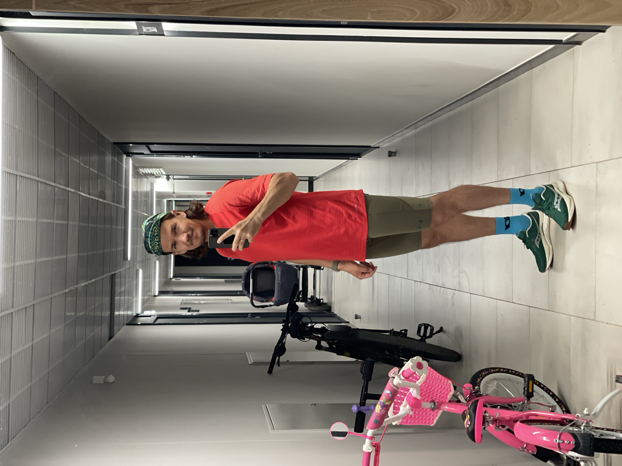 A man is captured in a mirror selfie, standing in what appears to be a well-lit indoor hallway or corridor. He has light brown hair, a slight beard, and a faint smile, holding a dark-colored smartphone to take the picture.
He is wearing a bright red short-sleeved t-shirt, olive green athletic shorts (with a small, indistinct logo, possibly Under Armour, on the left leg), and blue ankle socks that feature white lettering, possibly "DA". His footwear consists of dark green and white athletic shoes, which seem to have branding on the side and heel. A patterned green, white, and blue bandanna or cap, with barely discernible text that might read "GLADIAG", covers his head. A light-colored watch or fitness tracker is visible on his left wrist.
To the man's right (and left in the reflection), two bicycles are parked. The most prominent is a bright pink children's bicycle equipped with a white front basket and a small circular rearview mirror on the handlebars. Behind it, a darker-colored adult or larger bicycle is partially visible, with a dark bag or helmet resting near its seat.
The hallway features a light-colored tiled or concrete floor and light-colored walls. The wall on the left side (in the reflection) has a distinctive grid-like or ribbed paneling. The ceiling is also light-colored, fitted with modern linear LED light fixtures that provide bright illumination. The mirror itself has a wooden-toned frame or trim at the top. Further down the reflected hallway, doorways and a small white object, possibly a sensor, are visible on the wall. The overall setting suggests a modern interior, possibly a residential building or a facility with shared spaces.