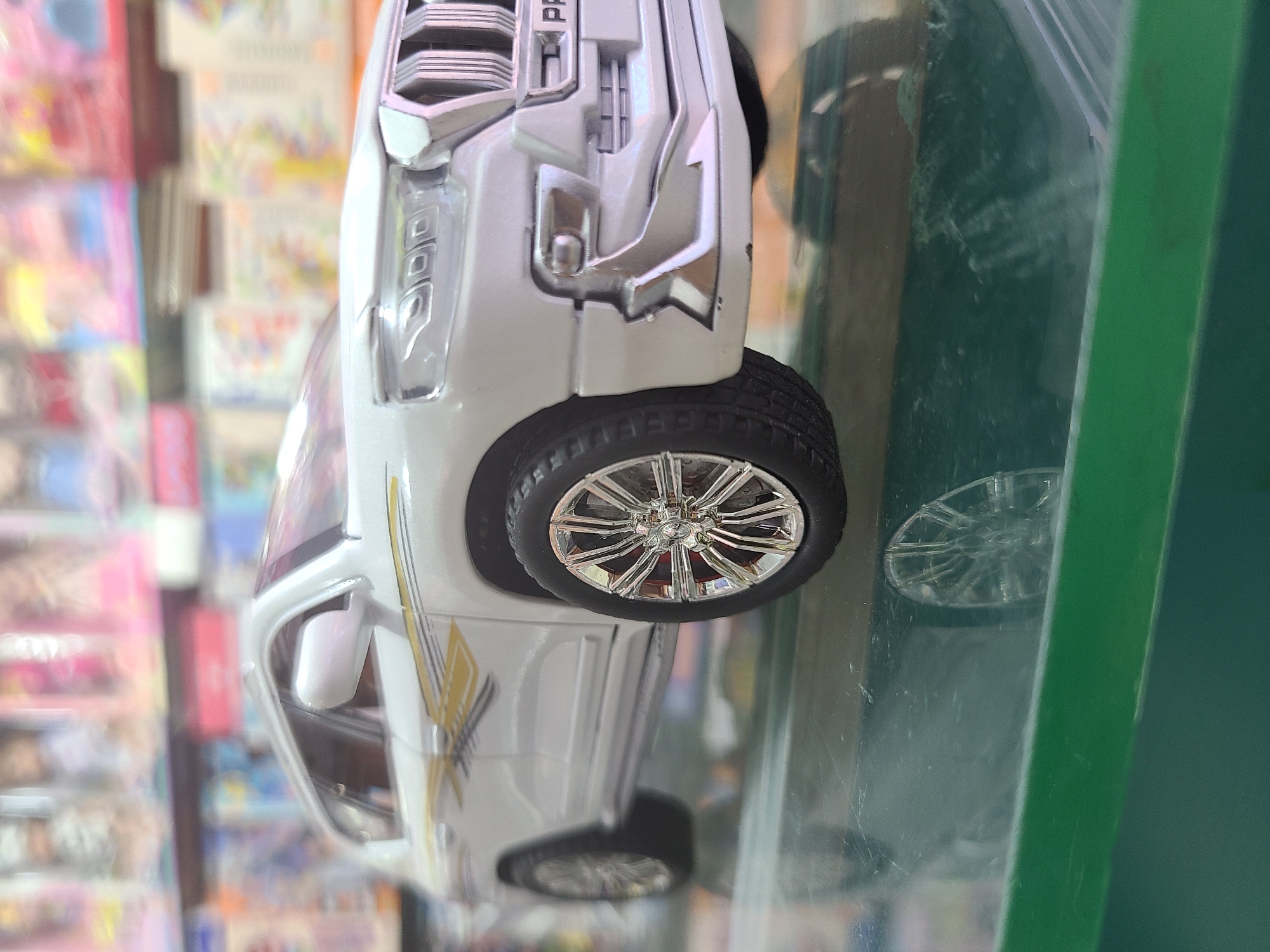 The image shows a white toy car inside a plastic container. The car has shiny, silver-colored wheels. A section of shelves with various colorful items is visible in the background. The setting is likely a store or home environment. The location is Quetta City Tehsil, Pakistan.