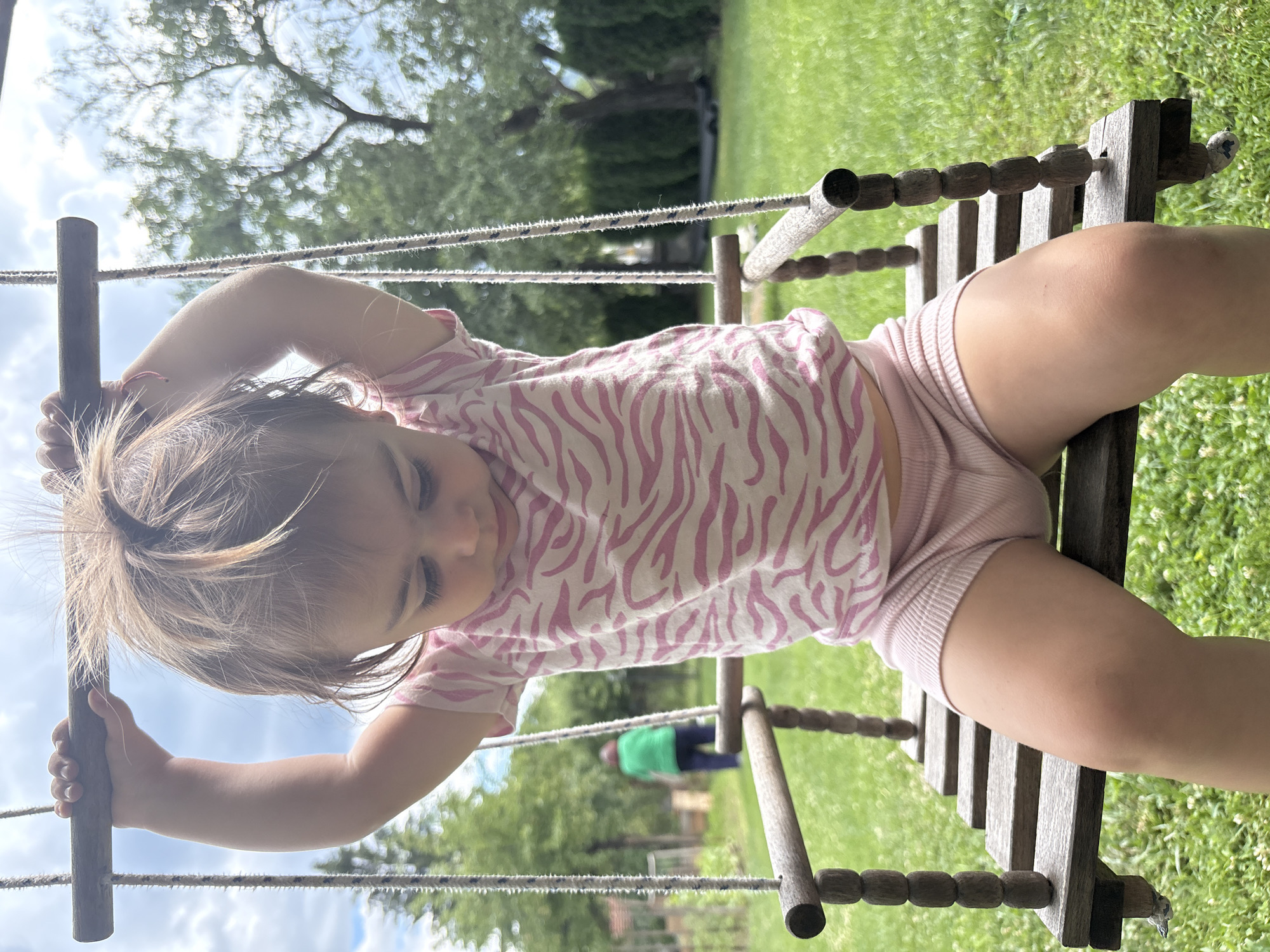 The media file shows a young child lying on a wooden swing, holding onto the wooden frame. The child is wearing a pink and white zebra-print t-shirt and pink shorts. The swing is suspended by ropes, and is outdoors in a grassy area with trees in the background. The sky is cloudy and bright, suggesting it is daytime. The child's eyes are closed, and they appear relaxed. In the background, another person wearing a green shirt and dark pants can be seen standing near a wooden structure. The overall scene suggests a casual, leisurely activity in a backyard or park setting. The location is identified as Oreshak, Bulgaria.