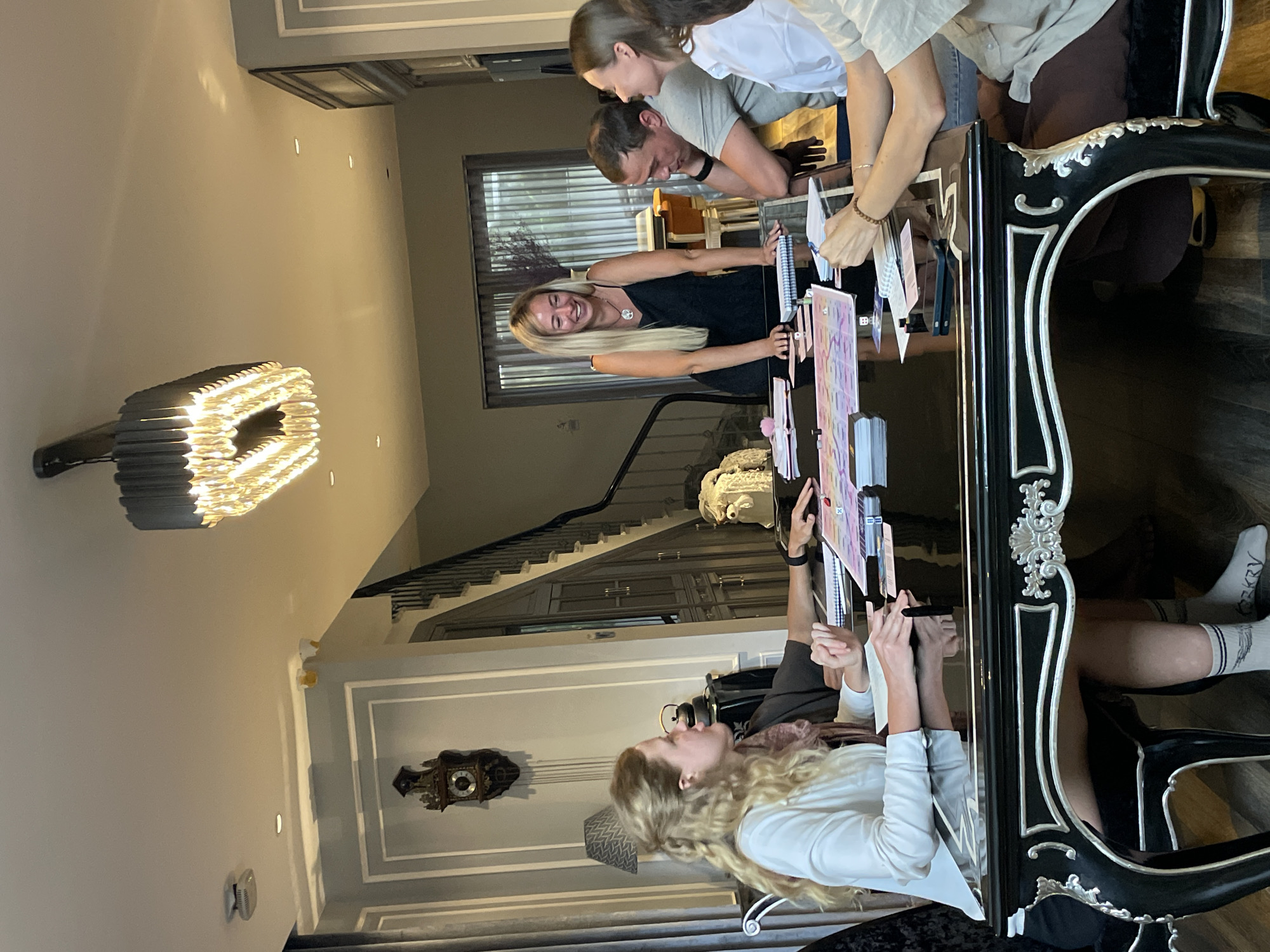 A group of five individuals are gathered around a glossy black table, engaged in what appears to be a board game or card game. The room is well-lit, with a modern chandelier hanging from the ceiling. To the left, a grandfather clock is mounted on the wall above a patterned lampshade. A staircase with dark railings and a dark wooden banister leads upstairs. The individuals are casually dressed and appear to be in good spirits, with one woman standing and laughing. The table is covered with game components, including cards, dice, and what looks like a game board. The flooring is dark wood. The scene suggests a social gathering in a home environment. The specific location within Xuan Truong - Da Lat, Vietnam cannot be determined from the visual information provided.