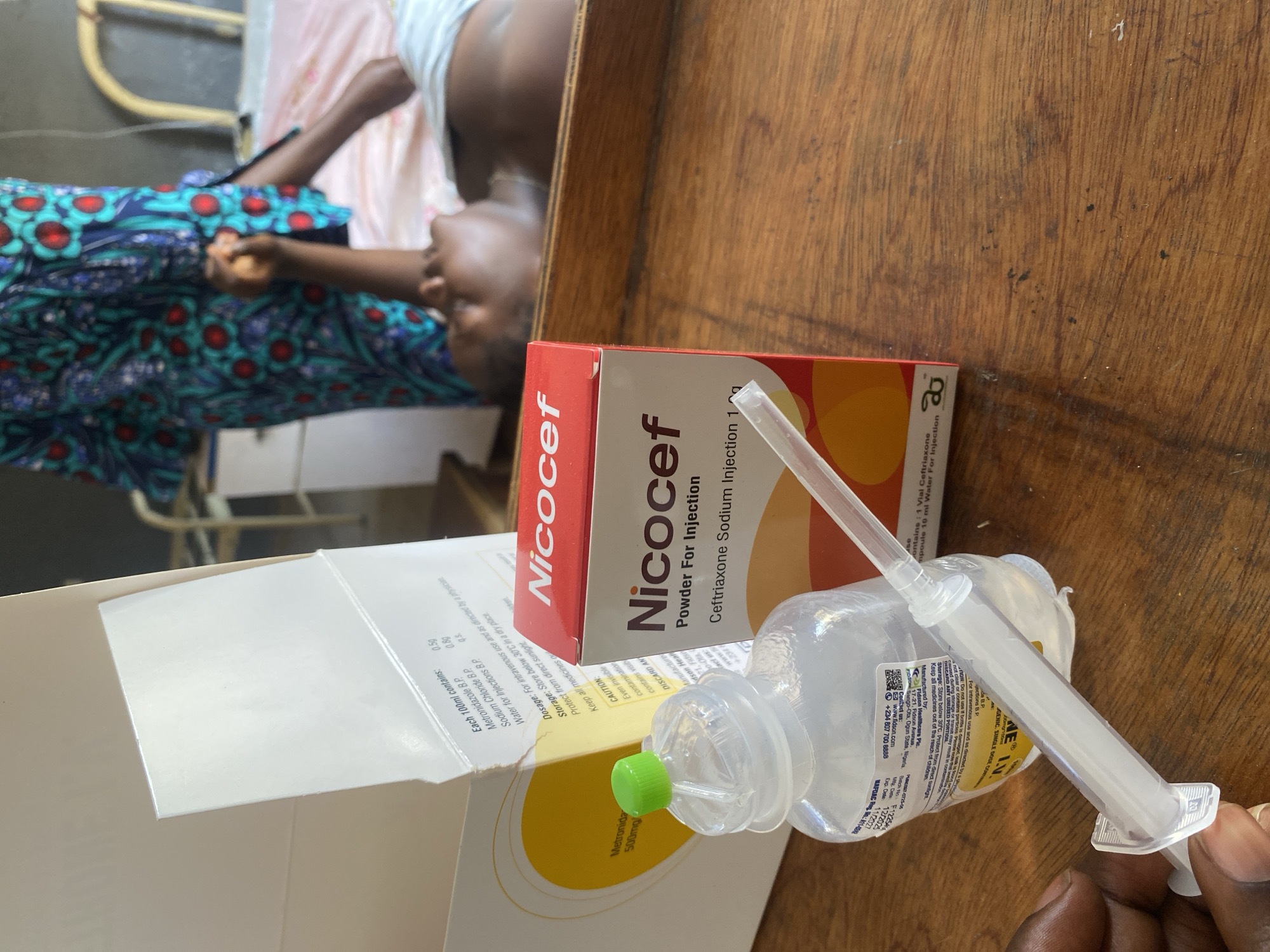 The image appears to be taken in a medical setting, possibly a hospital room, in Rido, Nigeria. A person is lying on a bed covered with patterned fabric. On a wooden surface next to the bed, there's medical equipment, including a box of Nicocef (Ceftriaxone Sodium Injection) powder, a vial labeled "Metronidazole 500mg," and a syringe. The medical items suggest treatment is being administered. The hand of a person can be seen holding the syringe.