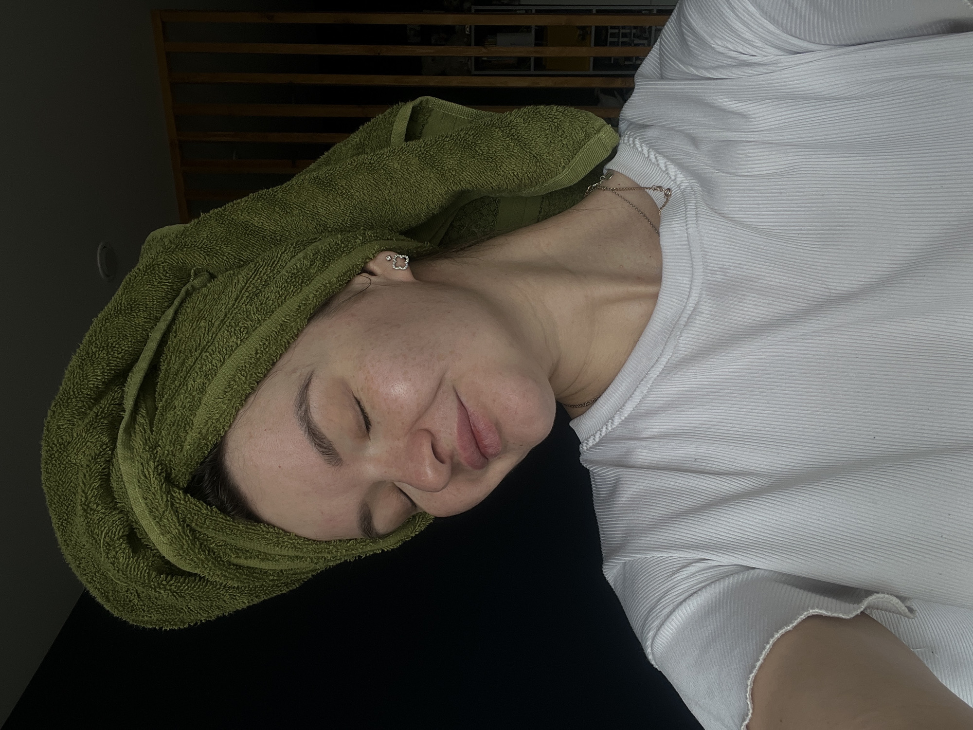 A woman with a green towel wrapped around her head is lying on a black surface, wearing a white ribbed shirt. She has a necklace and earrings. The location appears to be indoors, possibly in Kazan, Russia, based on unconfirmed contextual information (not visually apparent in the image). The woman's eyes are closed, and she has a slight smile.