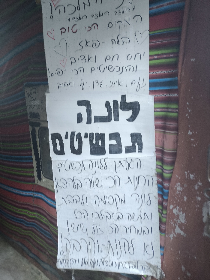 The image features two handwritten signs in Hebrew. The top sign contains endearments, including "the most awesome place" and a heart symbol, along with other phrases. The bottom sign prominently displays "Luna in the fields" in bold, above smaller, more densely written text. A "WG Termilos" logo is partially visible on a separate surface beside the signs.