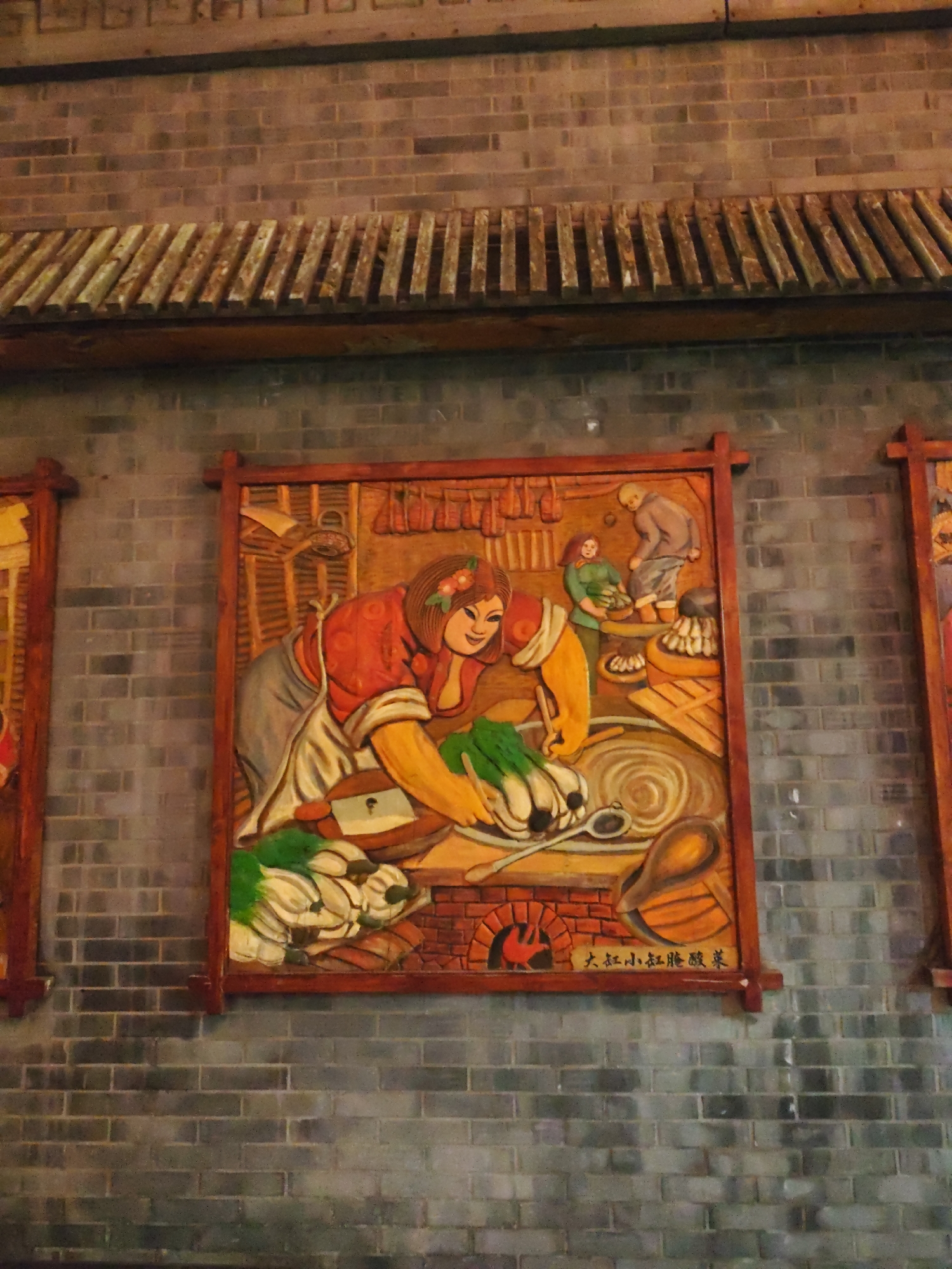 This image displays a carved wooden relief artwork depicting a scene of domestic activity, likely in a rural Chinese setting. The artwork is mounted on a brick wall, and the relief itself is framed by a wooden border. The central panel of the artwork features three figures. On the left, a woman with a red top and a white apron is shown chopping vegetables, which appear to be leeks or similar greens, in a large bowl. To her right, another woman in a green outfit is tending to a pot. Further back, a man in a grey garment is interacting with some produce, possibly potatoes or other root vegetables, which are piled in a basket. Behind the figures, elements of a kitchen environment are depicted, including what looks like dried meats hanging from the ceiling and a brick oven in the foreground. The artwork is signed with Chinese characters at the bottom right. To the left and right of the central panel, partially visible are other similar framed relief artworks. The overall impression is one of a traditional Chinese kitchen scene, possibly representing a community or family preparing food.
