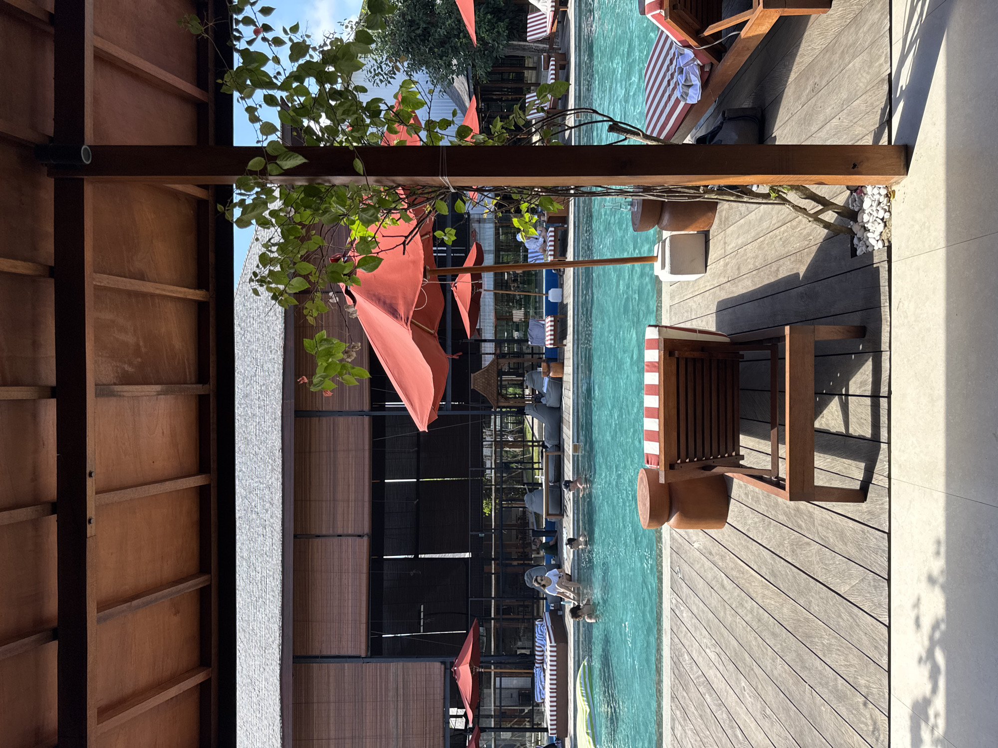 This photograph depicts a poolside setting. The scene includes people swimming, lounging in chairs, and interacting with the environment, which includes wooden decking, red umbrellas, and a building with blinds. The location is Kuta Selatan, Indonesia, evidenced by the distinctive architectural style and vegetation.