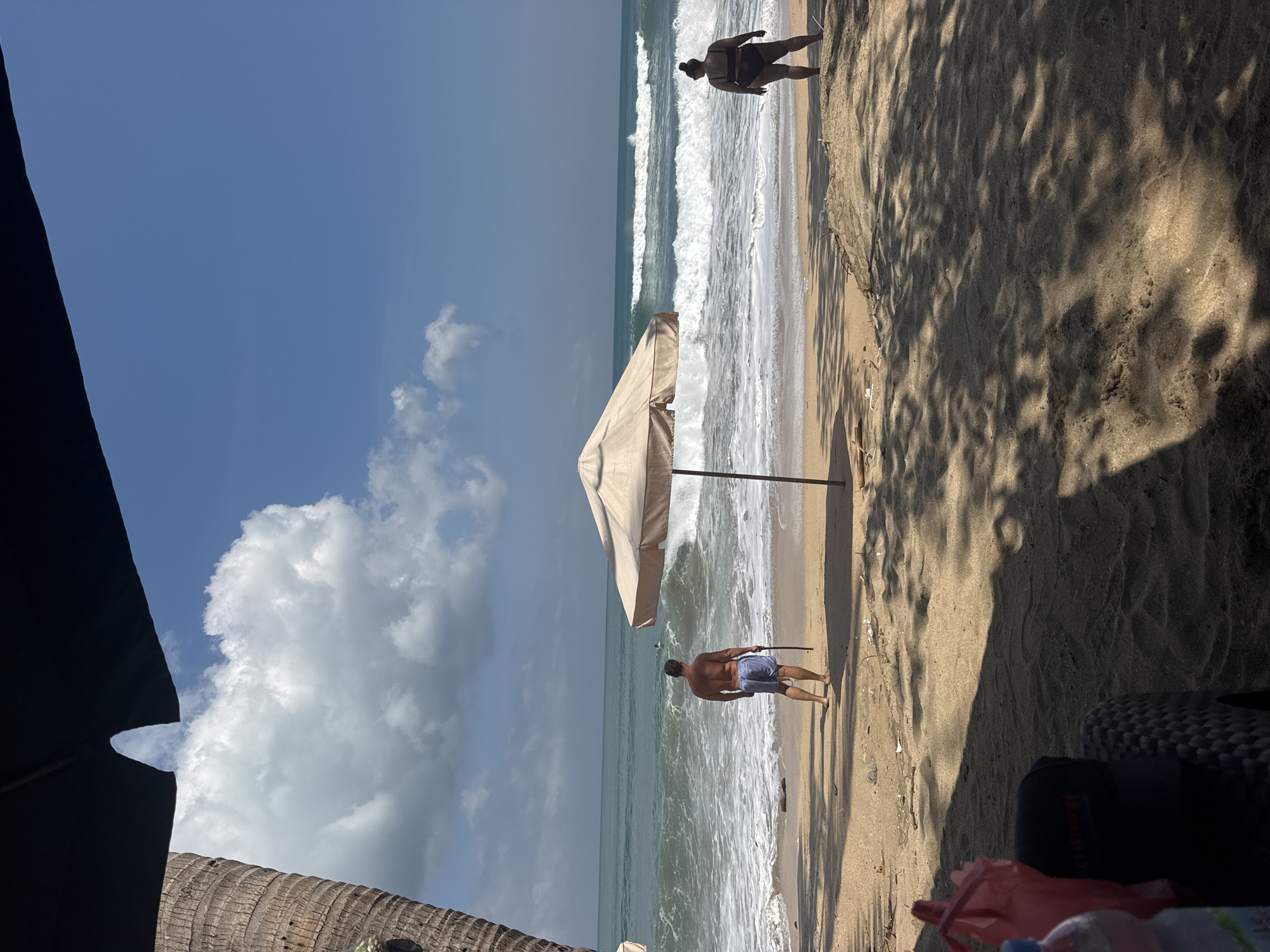 In the city of Kuta, Indonesia, a beach scene unfolds under a partly cloudy blue sky. The ocean exhibits moderate waves, with whitecaps breaking near the shore. Two individuals are present on the sandy beach. In the mid-ground, a man facing away from the camera stands with his back to the sea, wearing blue shorts and holding a stick. Further to the right, also facing the ocean, a woman stands wearing a dark bikini. A large, tan beach umbrella stands between them. The foreground is shaded by palm fronds, casting dappled shadows on the sand. Several items are visible in the shaded foreground, including a red plastic bag and a dark baseball cap on top of a woven object. To the left, the trunk of a palm tree is visible.