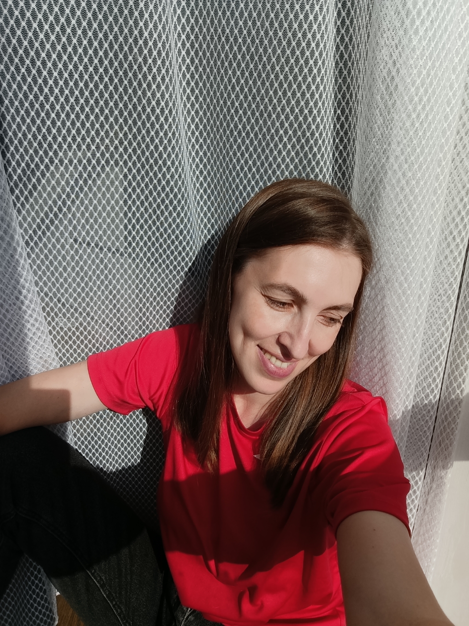 In the city of Chaikovsky, Russia, a woman with shoulder-length brown hair smiles while looking down. She is wearing a red t-shirt and dark pants. The woman is positioned in front of a white, net-like curtain that is casting shadows. Sunlight streams in from the right, illuminating her face and casting a reddish hue on her clothing. The background suggests an indoor setting, possibly a room with a window.