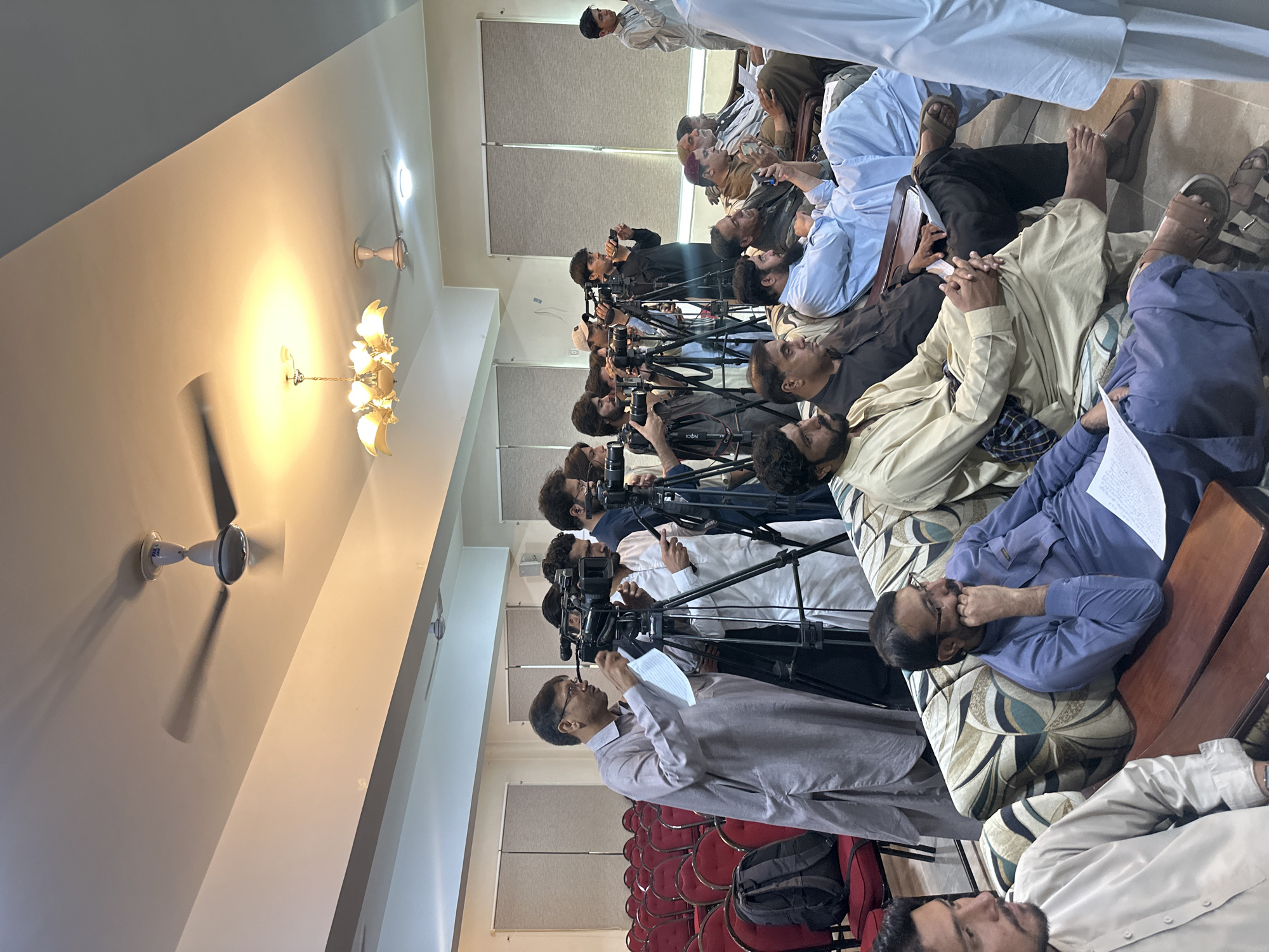 A room in Quetta City Tehsil, Pakistan contains a group of men, a row of cameras on tripods, and a series of horizontal window blinds. The men are mostly seated on the floor on cushions or mats. Many of the men are operating the cameras, which are pointed towards the other end of the room, but some are simply seated or reclining. A series of red chairs are visible. The room has a chandelier and ceiling fans.