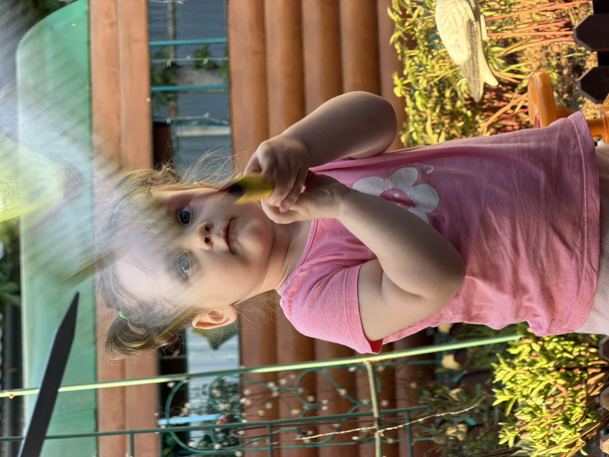 A toddler stands in a garden in what might be Chaikovsky, Russia, wearing a pink t-shirt with a white flower design. She holds a yellow object in her hands, looking at the camera. A green and brown wooden structure is behind her, with metal fencing and lush greenery visible in the background. A possible reflection or blurred object obscures the left side of the frame.