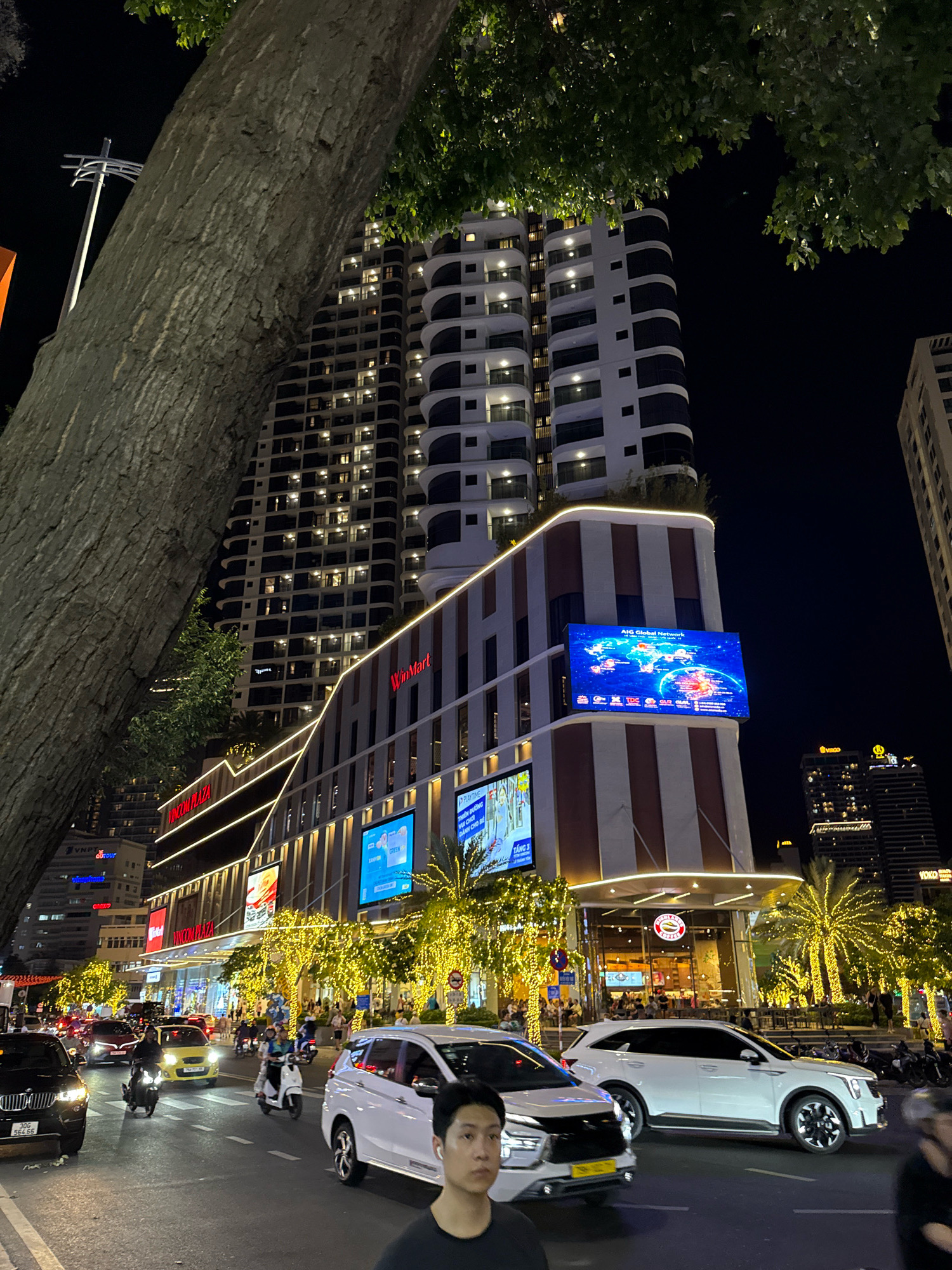 This is a nighttime street-level view in Nha Trang, Vietnam, focusing on a modern building complex adorned with bright lights. A large tree trunk dominates the left foreground, partially obscuring a tall, multi-story building with numerous illuminated windows and a curved architectural style. Below this, a section of the building features red and white striped columns and a prominent "WinMart" sign. A large, blue LED screen displays a world map with glowing points. In front of the building, palm trees wrapped in yellow lights line the sidewalk. The street is busy with traffic, including cars and motorcycles. A white SUV is stopped in the foreground, and a black BMW is visible on the left. Several people are seen on the street, some on motorcycles and others walking. The overall atmosphere is vibrant and illuminated.