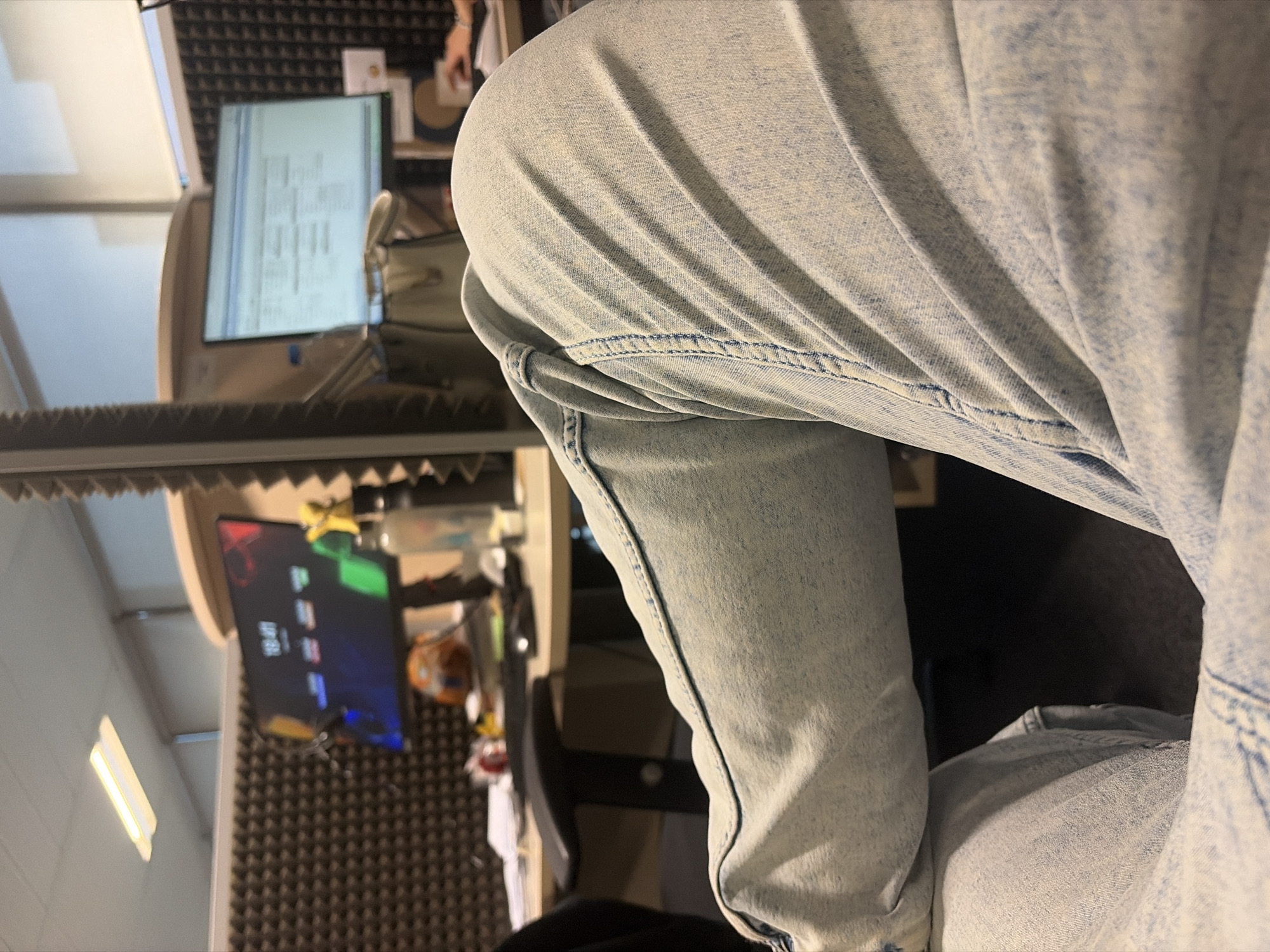 A medium shot depicts a person wearing light-wash denim pants, possibly jeans, from the waist up. The subject is seated. In the blurred background, a desk with computer monitors and what appears to be soundproofing material is visible, suggesting an office or studio environment. There is a lighting fixture on the left. The exact location within Sofia, Bulgaria, cannot be determined from the provided image.