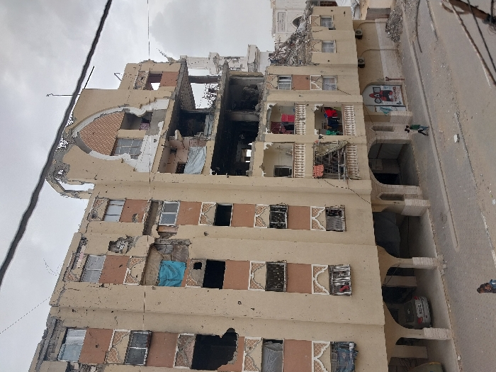 A multi-story residential building in Khan Yunis, Palestinian Territory, displays extensive structural damage under an overcast sky. The approximately seven-story, beige-colored concrete structure exhibits large sections of its facade blown out, revealing internal rooms and exposed construction elements. Numerous windows are shattered or entirely absent, with some openings covered by blue and white tarpaulins. The upper right section features a heavily damaged arched architectural element, exposing internal brickwork. The building's exterior is marked by multiple impact points consistent with bullet or shrapnel strikes. Several mid-level balconies remain partially intact, with various items of laundry hanging. The ground level incorporates arched open bays; one on the left contains a parked silver sedan. Graffiti, including a red stylized figure and Arabic script, is visible on a ground-floor wall. Two individuals are walking on the sidewalk in the foreground, and one additional individual is visible further down the street. Piles of rubble are accumulated at the building's base and along the adjacent street. In the background to the right, a damaged or incomplete white building, possibly a mosque with an arched entrance, stands partially obscured by debris.