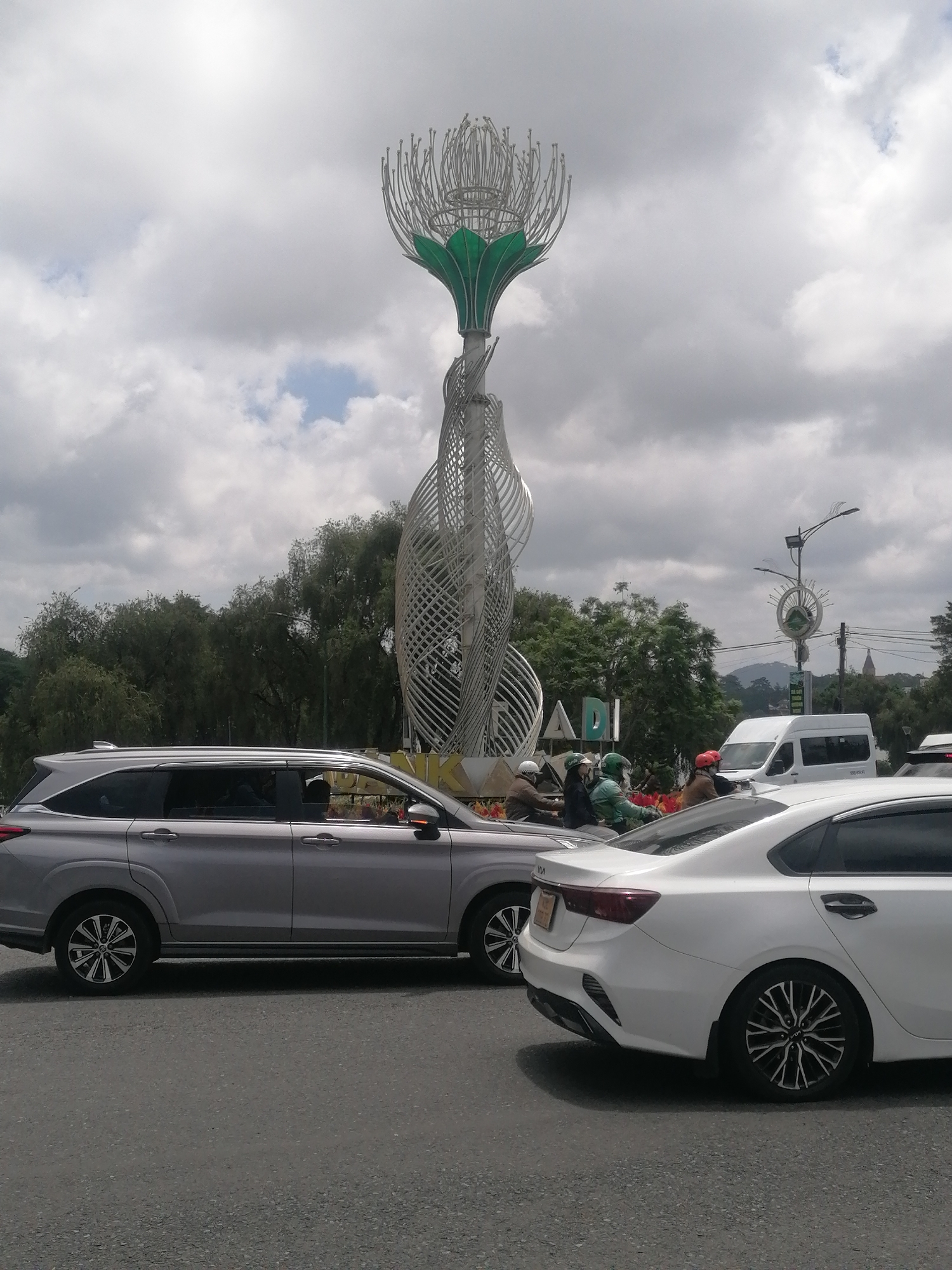 The image depicts a street scene in Da Lat, Vietnam, dominated by a large, modern metal sculpture resembling a stylized flower. The sculpture is tall and intricately designed with swirling metallic elements and green leaf-like accents at its base. In the foreground, two cars are visible on the asphalt road. On the left is a silver SUV, and to its right is a white sedan. Several people on motorbikes are also present, some wearing helmets, suggesting typical city traffic. The background features a line of lush green trees, a white van, and a streetlamp with a circular design. The sky is overcast with scattered clouds. The overall environment suggests an urban setting with some natural elements.