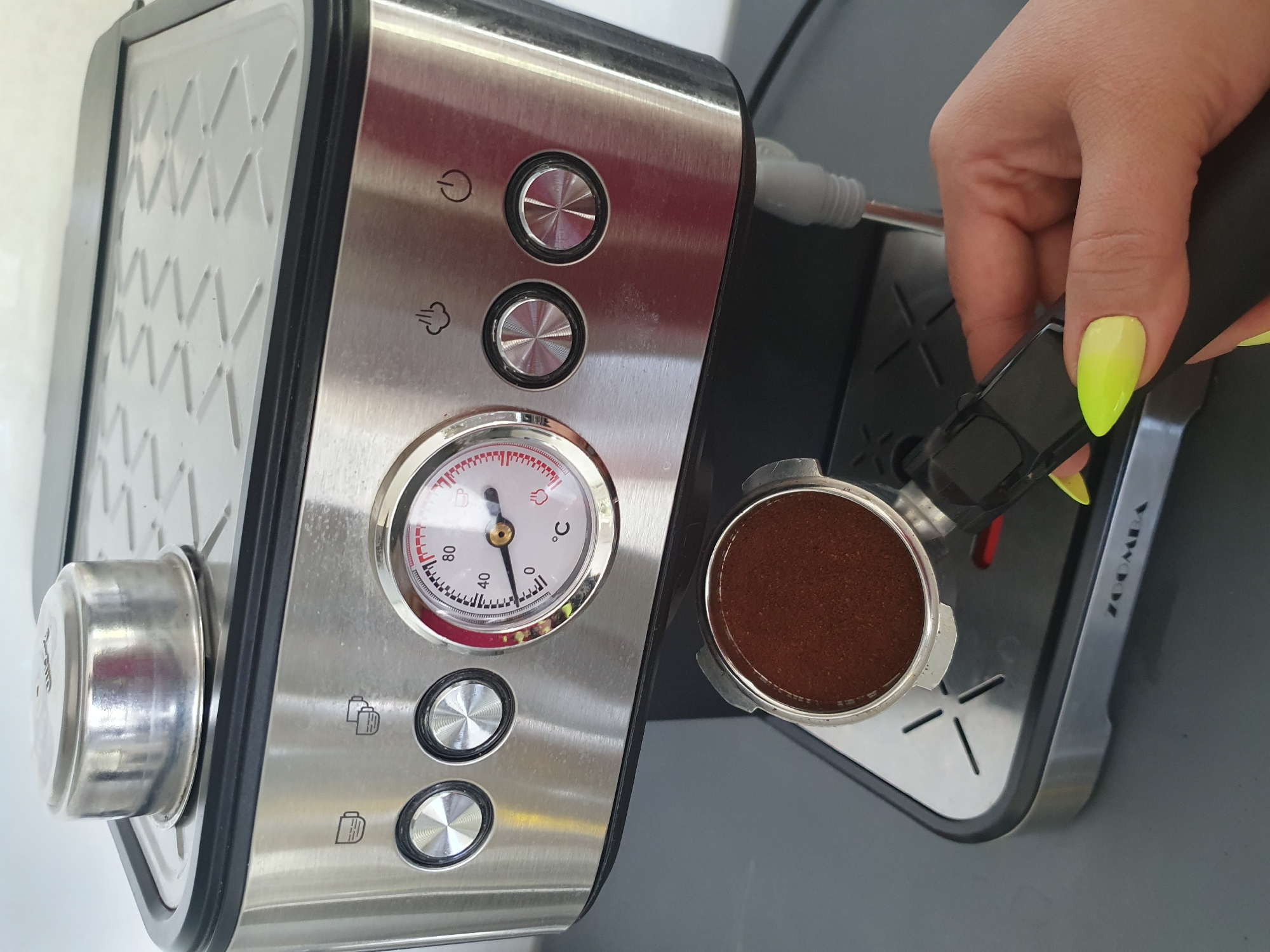 A person's hand with neon yellow manicured fingernails is holding a portafilter filled with dark brown coffee grounds. The portafilter is positioned over a drip tray on a stainless steel espresso machine. The espresso machine has a silver cup warmer on top, a dial with temperature markings from 0 to 80 degrees Celsius, and several control buttons with icons for steam, a power symbol, and cups. The machine is sitting on a grey surface, likely a countertop. The background is out of focus but appears to be an indoor setting.