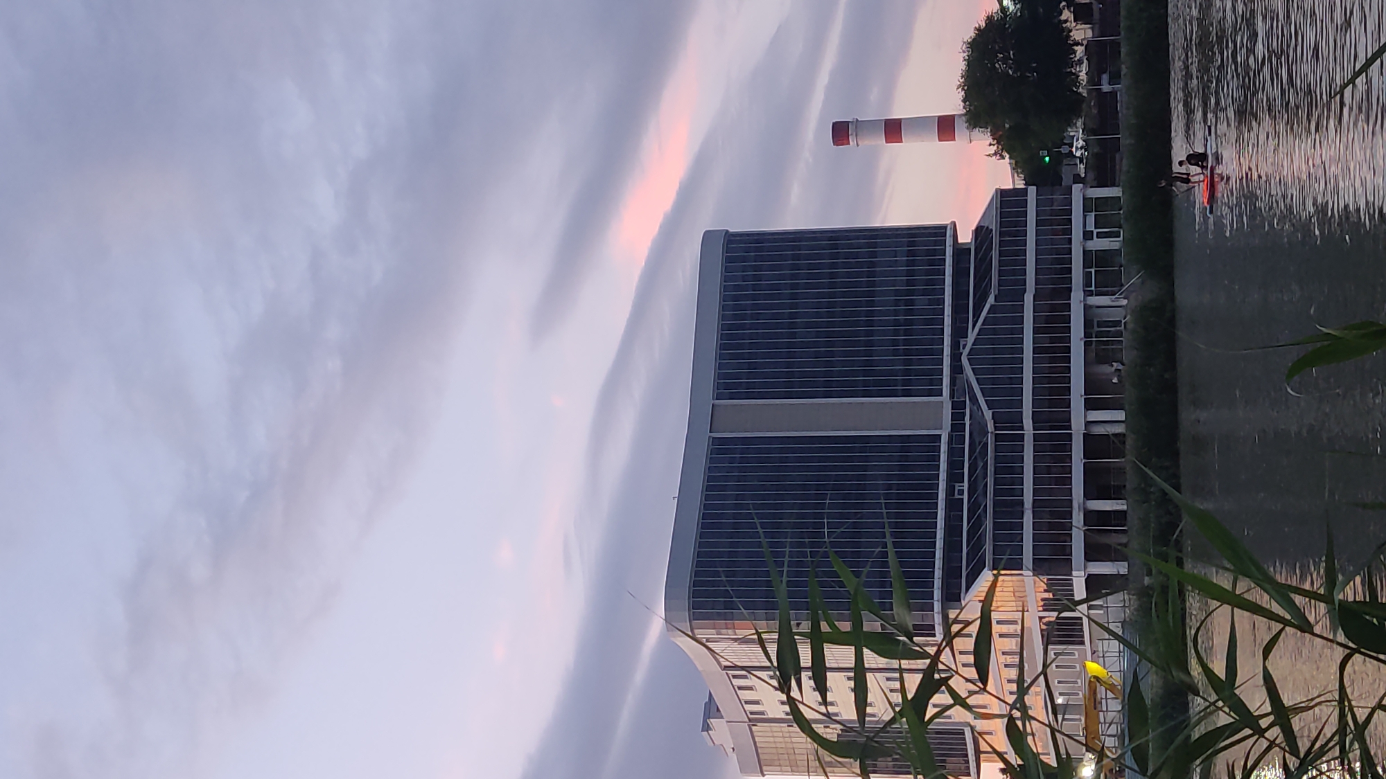 The image depicts an evening scene by a body of water, likely a canal or river, in Krasnodar, Russia. A modern, multi-story building with a dark, grid-like facade dominates the mid-ground, reflecting the dusky sky. To the left of the building, a tall, cylindrical smokestack with red and white stripes stands out against the sky. The sky itself is filled with wispy, elongated clouds, some tinged with pink from the setting sun, suggesting it is either dawn or dusk.
In the foreground, tall, green reeds or grasses partially obscure the view, adding a natural element to the urban landscape. On the water, two figures can be seen on a small illuminated raft or boat, engaged in an activity that is not clearly discernible but appears to involve some form of recreation. The water is rippled, reflecting the muted colors of the sky and the surrounding structures. The overall mood is tranquil and somewhat atmospheric, with the fading light casting a serene glow over the scene. There is no visible text in the image.
