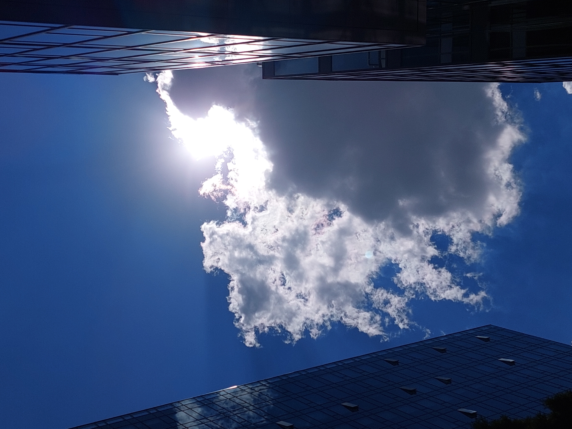 A low-angle, full shot captures the sky above a city, framed by the dark glass facades of two modern buildings. On the left, a building with a grid pattern of windows stretches upwards, reflecting the bright blue sky and scattered white clouds. On the right, a taller building, also clad in dark glass with a grid of vertical lines, dominates the right side of the frame. The sun, obscured by a large, fluffy white cloud, shines intensely in the upper center, casting bright rays and creating a lens flare effect. The cloud has both brightly illuminated sections and darker, shadowed areas, adding depth and contrast to the sky. The sky is a vibrant blue, with smaller, wispy clouds visible in some areas.