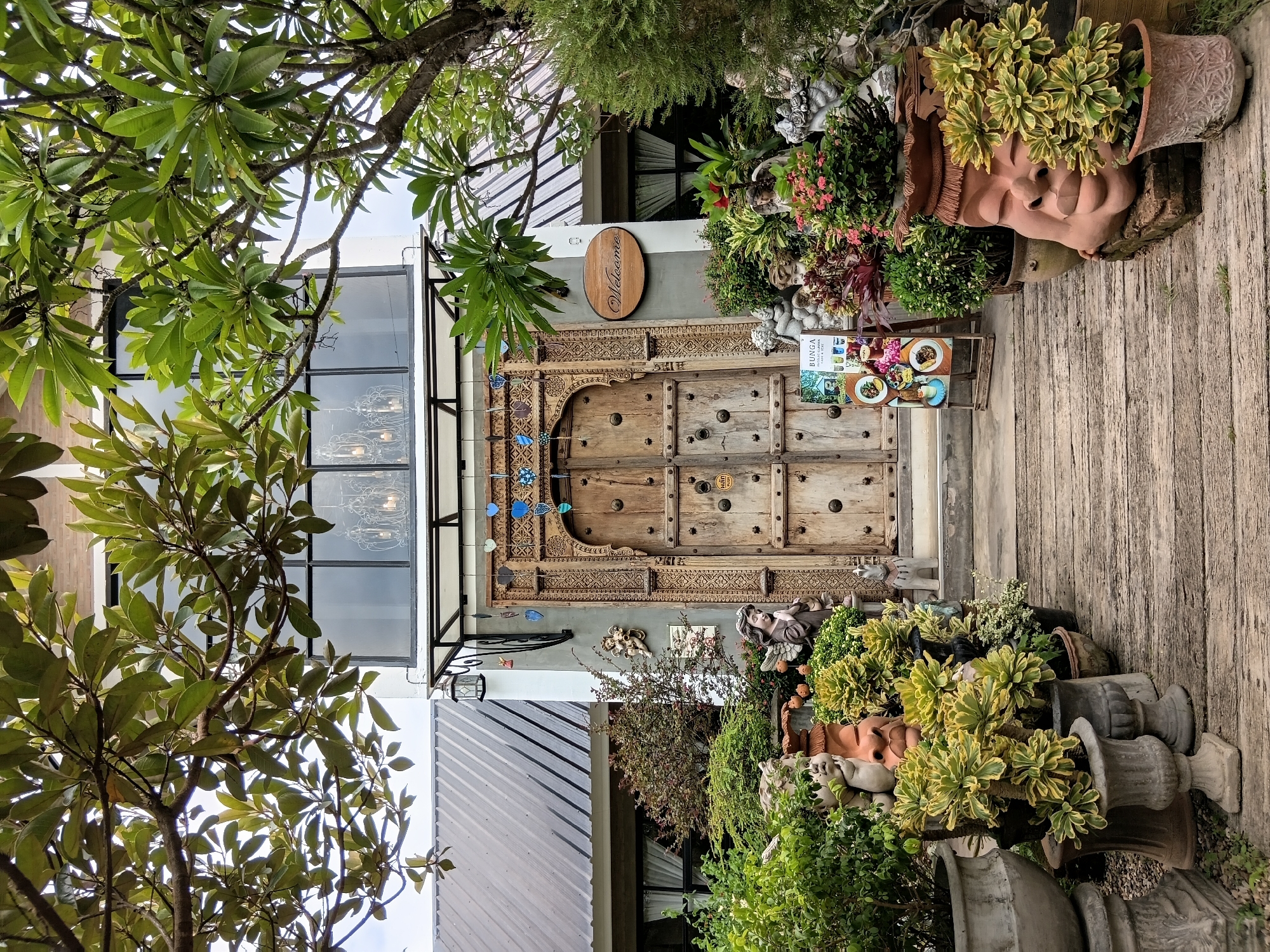 The image depicts the entrance to a building in Ban Tong, Thailand. The entrance features a large, ornate wooden door with decorative carvings and metal studs. To the left of the door, there is a wooden sign with the word "Welcome" in cursive script. The facade of the building is white with dark-framed windows. Above the entrance, a balcony area is visible with large windows and a chandelier inside. Lush greenery and potted plants surround the entrance, with various garden statues, including cherubs and a large face sculpture, placed amongst the foliage. A wooden pathway leads to the door. The overall impression is one of a welcoming and intricately decorated establishment.