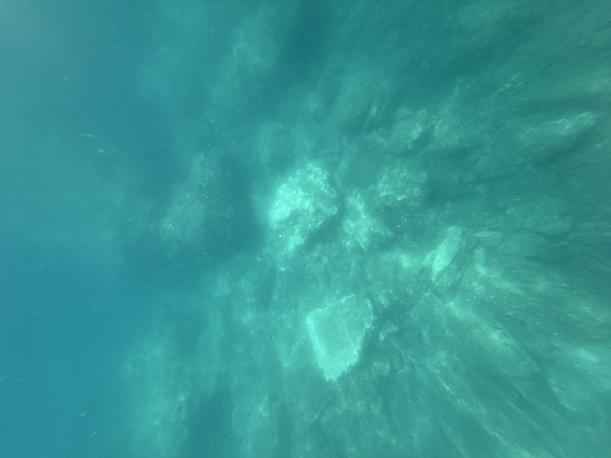 The image depicts an underwater environment in an unknown location, city of Unknown. The water is clear and exhibits a dominant turquoise hue. The seabed is composed of numerous irregular rock formations of varying sizes, some appearing lighter due to illumination or surface characteristics. Fine particulate matter is discernible suspended throughout the water column. The perspective is an angled view looking downwards onto the rocky bottom, with ambient light filtering from above. No people, animals, or specific man-made objects are visible.
