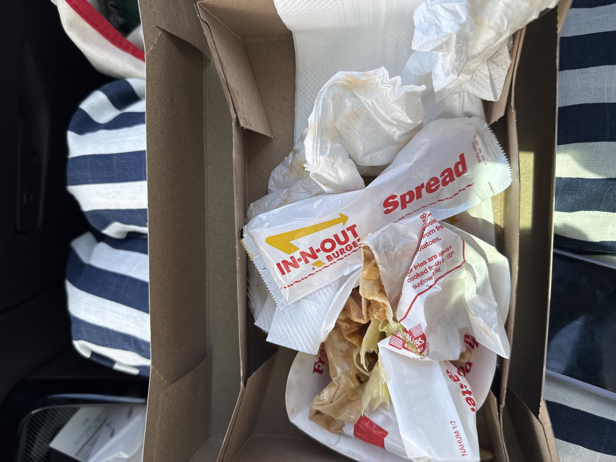 A high-angle close-up shows a partially consumed meal from the fast-food chain In-N-Out Burger inside a cardboard box. The box appears to be discarded and contains used white napkins, a packet of "Spread" sauce, a wrapper for the burger with the In-N-Out logo, and remnants of fries and other food debris. The box is situated on what appears to be a car seat, with a blue and white striped cloth partially visible beside it. The overall impression is that the meal has been eaten and the remnants are being discarded. The image does not contain information to conclude the location of Desert Hot Springs, United States of America.
