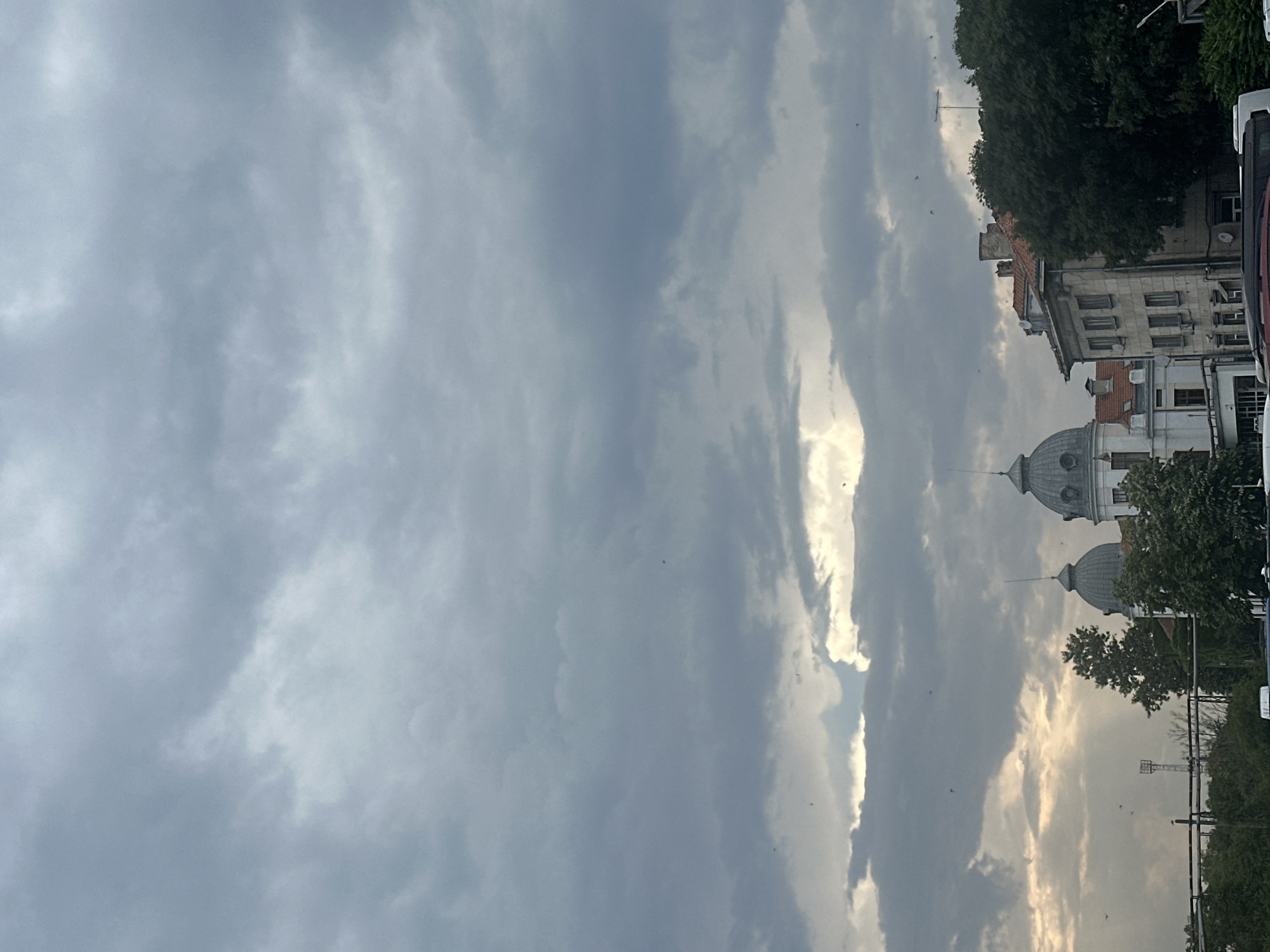 The image captures a cloudy sky over a cityscape. Several buildings with distinctive domed roofs are visible, suggesting a potentially historical or architecturally significant area. Trees and greenery are interspersed with the urban structures. The sky is predominantly gray with patches of lighter clouds, and some sunlight appears to be breaking through in the distance. A few small, dark shapes, possibly birds, are scattered across the sky. The overall atmosphere is overcast, and the scene is static.