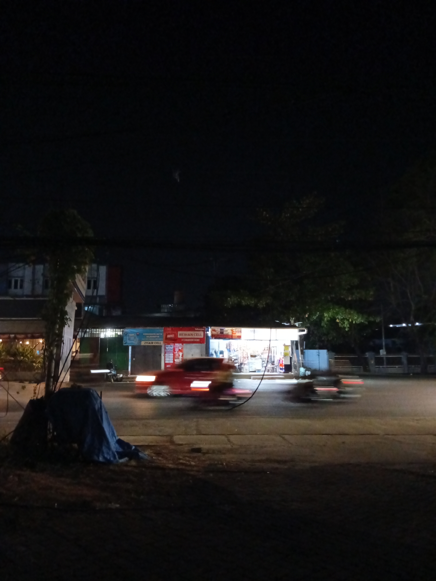 The image captures a night scene on a street in Makassar, Indonesia, likely near a commercial area given the illuminated storefronts. In the foreground, a dark, tarp-covered object, possibly temporary shelter or stored material, rests on the ground. The middle ground features a road with several vehicles captured in motion blur, indicating low light conditions and movement. A prominent red vehicle with illuminated headlights is moving from left to right. Further back, a brightly lit storefront displays signage, including "REIHAN CELL" and "MINI ATM," suggesting a small business or convenience store. The background reveals a building with windows and trees, contributing to the urban night atmosphere. The overall scene depicts a typical street view in Makassar at night, characterized by the interplay of artificial light from businesses and the motion of vehicles.