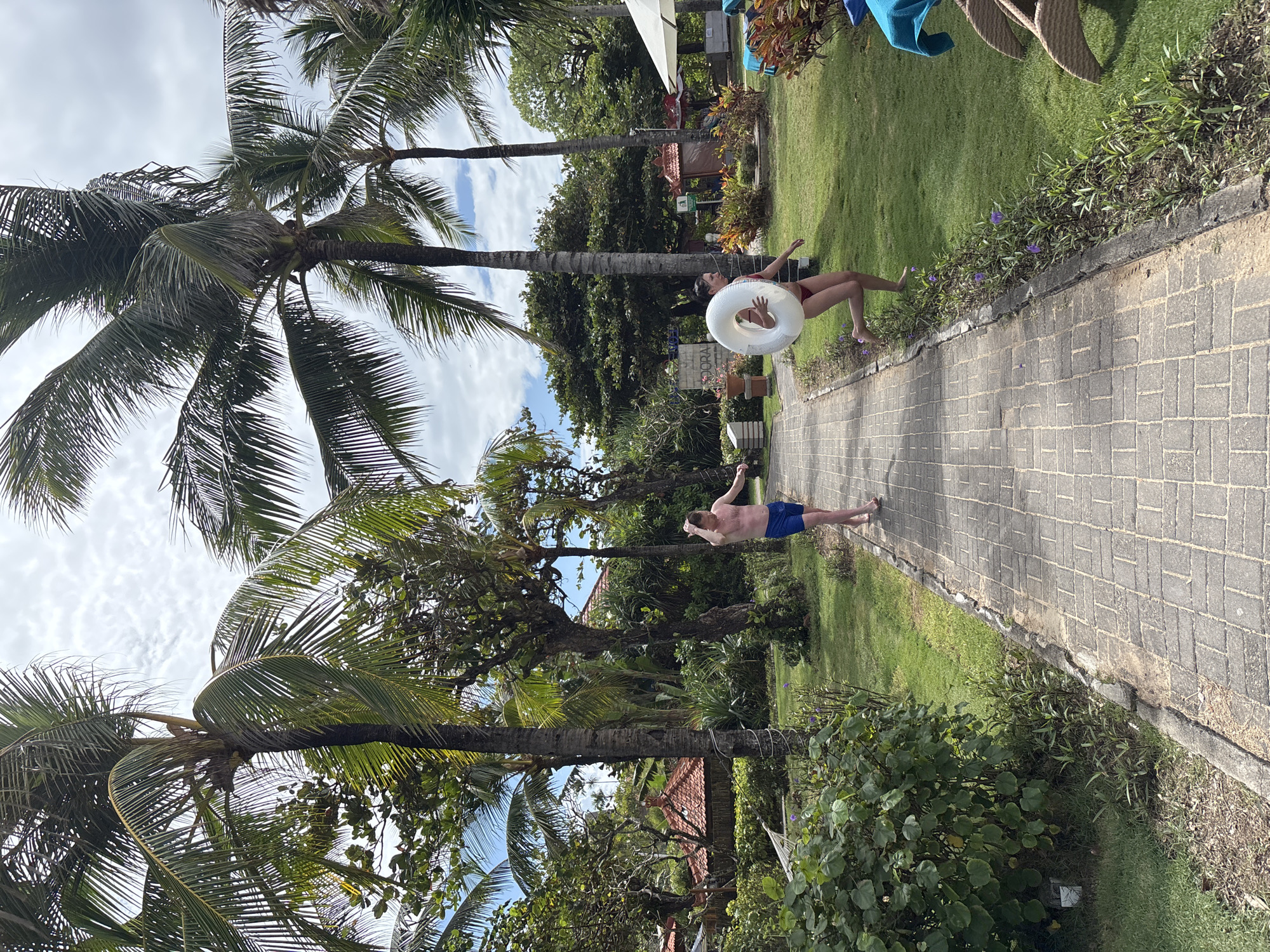 In Kuta Selatan, Indonesia, two individuals are captured in a candid moment on a paved pathway lined with lush green grass and tropical foliage. A man, wearing blue swim trunks, walks towards the camera with his right hand raised to his head, appearing relaxed. To his right, a woman, clad in a red bikini, runs away from the camera, holding a white inflatable ring. The pathway itself is constructed from interlocking rectangular bricks, bordered by a strip of small purple flowers and verdant plants. Beyond the pathway, palm trees with their distinctive fronds tower over the scene, with the sky peeking through in scattered clouds. In the background, traditional Indonesian-style roofs with terracotta tiles are visible, suggesting a resort or residential area. The overall atmosphere suggests a leisurely day in a tropical vacation setting.