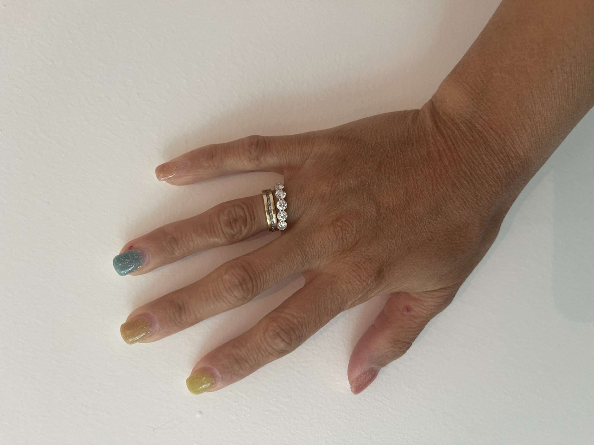A person's hand is shown with multi-colored nail polish and two rings on the ring finger, resting against a white wall. The hand has a medium skin tone and appears to be an adult's. The nail polish colors are yellow, teal, gold, and a peachy nude. One ring is a plain gold band, and the other appears to be a diamond band. The white wall is smooth with subtle texture. The scene is devoid of any urban indicators. This image cannot be geolocated to Ornek, Kyrgyzstan.