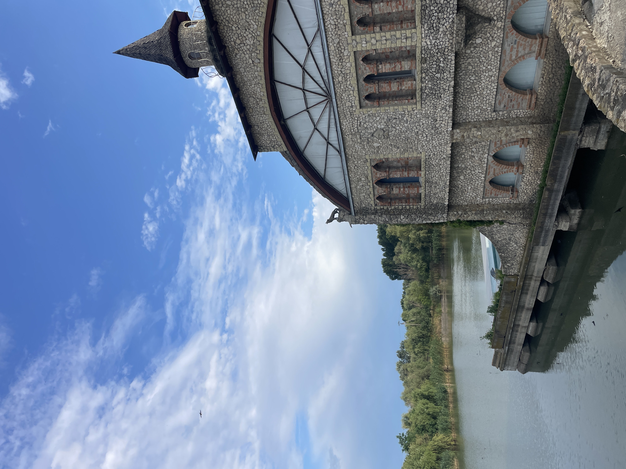 The image shows a stone building with a conical tower next to a body of water. The building has arched windows and a decorative stone wolf figure on its roof. The water is calm and reflects the building and the surrounding trees. The sky is blue with scattered clouds. There is a small airplane visible in the sky. The location is identified as Colibași, Moldova.