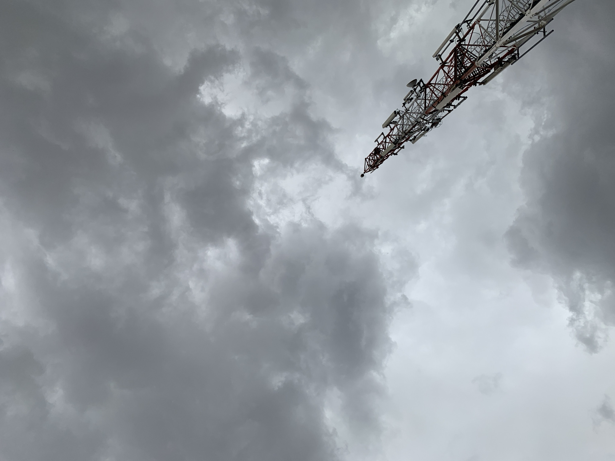 The image shows a communications tower against a cloudy sky. The tower is constructed with a lattice framework, painted white and red, and has various antennae attached to it. The clouds are dense and gray, indicating a potentially stormy weather. The scene is located in Rafin Par, Nigeria.