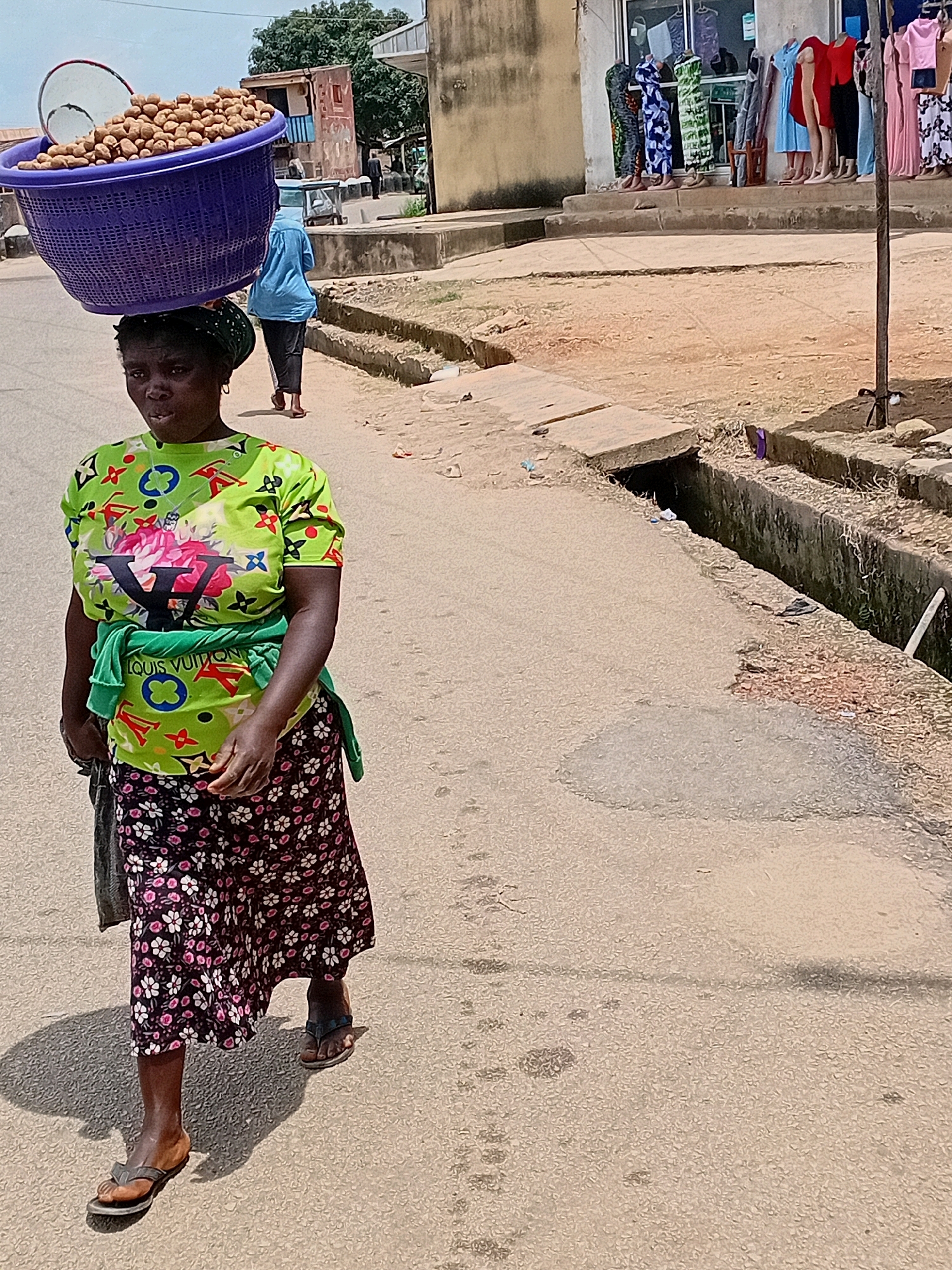A woman walks along a street in Jos, Nigeria, carrying a large basket of yams on her head. The woman is wearing a green t-shirt with a Louis Vuitton logo and floral print, a floral skirt, and sandals. She is also wearing a green headscarf. Behind her, another person walks away from the camera, wearing a blue shirt and dark pants. The street is paved with asphalt and has a gutter on the right side. In the background, there are buildings, including a shop with clothes displayed on mannequins. The sky is partly cloudy.
