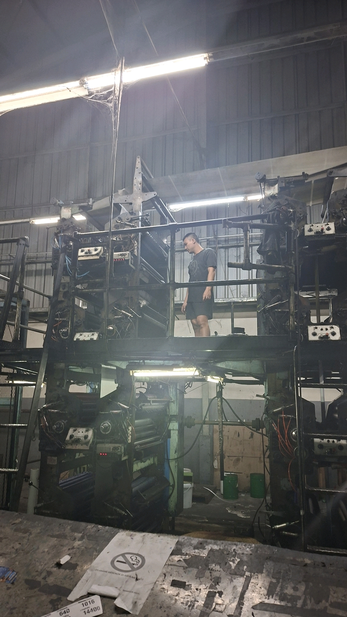 The image depicts a man standing on a raised platform within a large industrial space, likely a printing facility, given the presence of complex machinery resembling printing presses. The environment appears somewhat aged and dusty, with visible cobwebs hanging from the ceiling in the upper left quadrant. The walls are constructed of corrugated metal, and fluorescent lighting illuminates the scene. The floor is concrete and shows signs of wear and debris, including discarded paper or plastic sheets, one of which displays a "no smoking" symbol and text in Indonesian. The man, wearing a dark t-shirt and shorts, is looking down and to his left, seemingly observing the machinery. Two green buckets and a white bin are visible on the floor in the background. The overall impression is of an operational but possibly older industrial setting in Palembang, Indonesia.