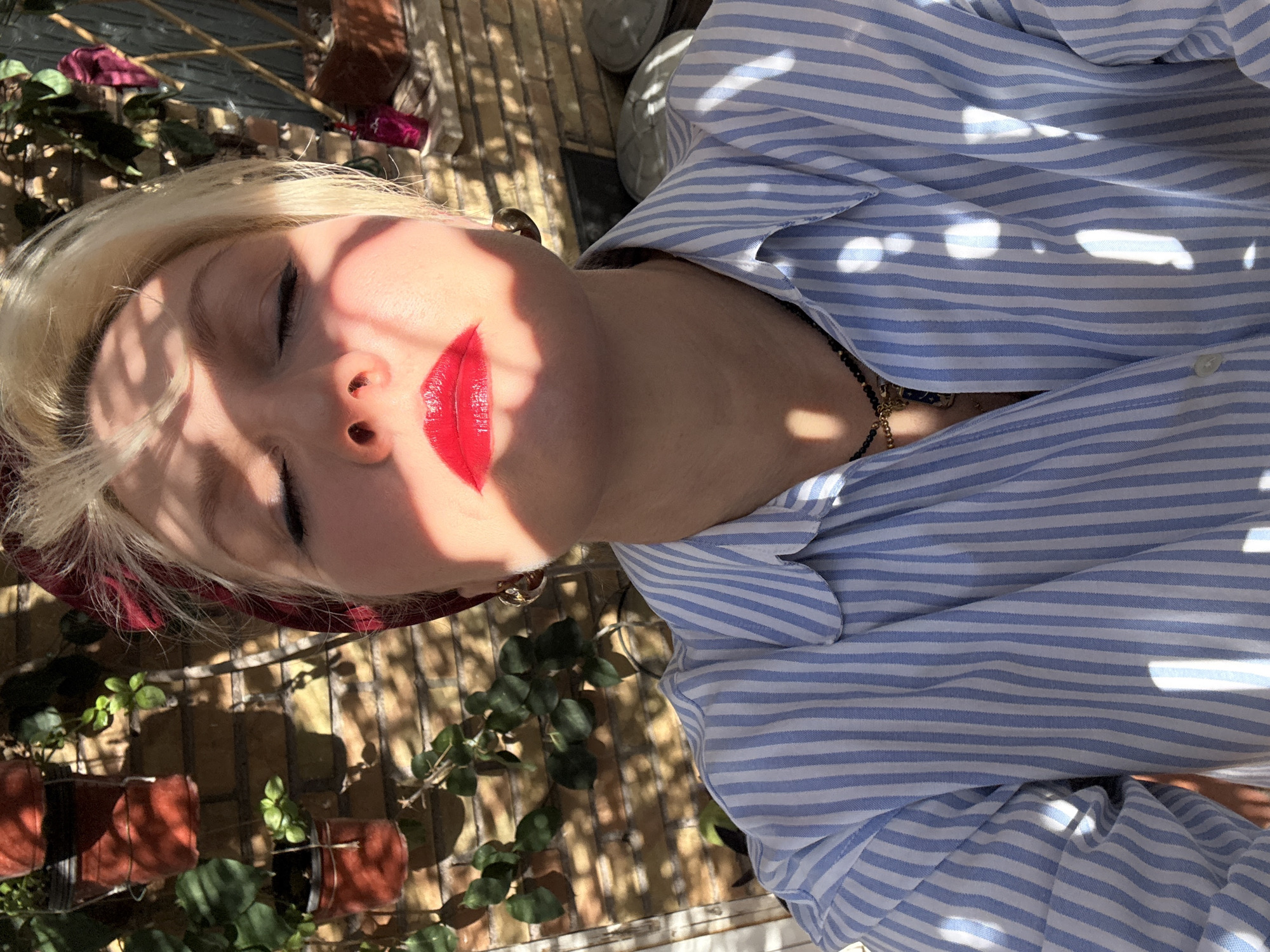 A woman with pale skin and blond hair is lying down, wearing a blue and white striped shirt. She has her eyes closed and is wearing red lipstick. The scene is outdoors, with plants in terracotta pots visible in the background. There are shadows from sunlight dappling the scene. A brick wall can be seen, along with what appears to be a wooden trellis. A burgundy headscarf can be seen on the woman's head. It is difficult to determine the exact location without further context, but the presence of terracotta pots, brick, plants, and sunlight suggest a possible balcony or courtyard in Rome, Italy.