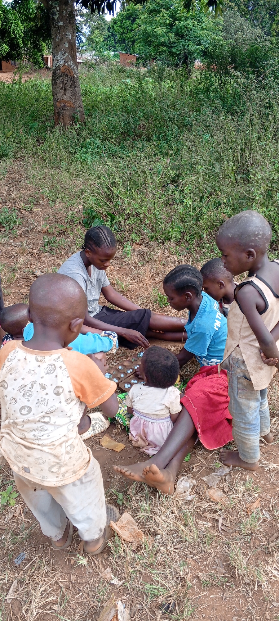 In Ngalula, Democratic Republic of the Congo, a group of children play a game outdoors on a dirt ground partially covered with dry grass and sparse greenery. The children are of African descent, with varying ages and clothing. One child is seated on the ground wearing a red skirt, while another wears a light-colored dress. An adult is seated with the children, wearing a grey t-shirt. A wooden game board with marbles sits in front of them. A young child stands in the foreground, wearing a patterned shirt and pants. A tree is visible in the background amongst other greenery.