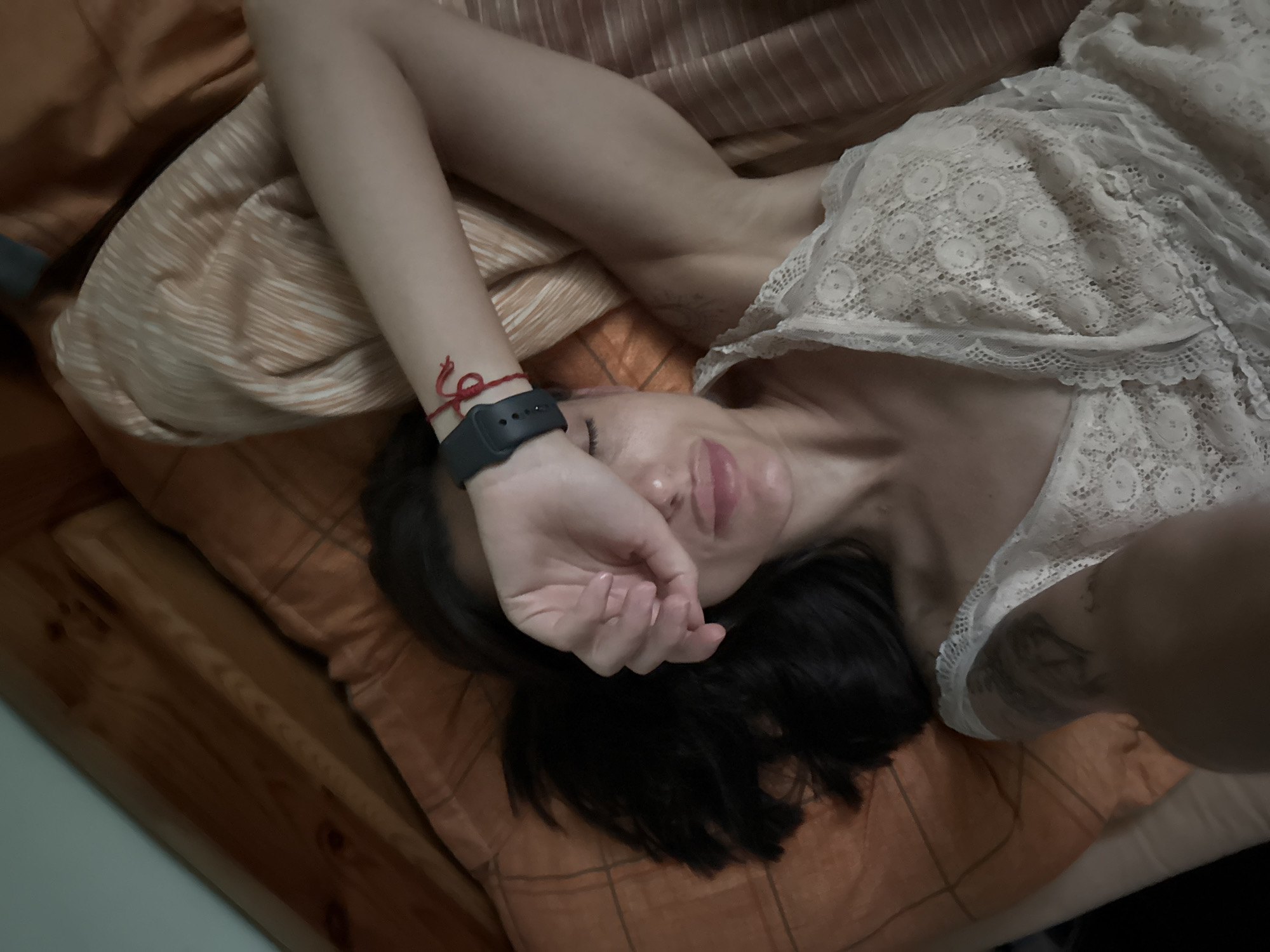 The image shows a top-down view of a woman lying on a bed. She is wearing a white lace top and has her left arm raised, covering her eyes. A black smartwatch is visible on her left wrist, along with a red string bracelet. Her hair is dark and spread out on the orange and white plaid bedding. The background consists of a wooden headboard and a light blue wall. The scene is indoors, likely a bedroom. The location in the city of Pleven, Bulgaria is not determinable from the image itself.