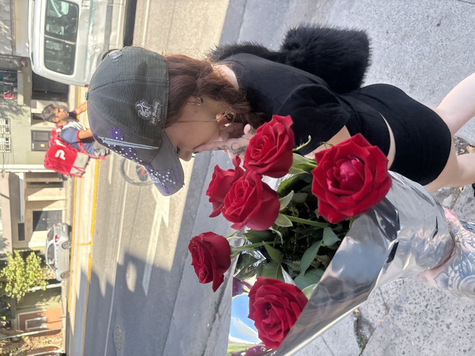 A woman wearing a black outfit, fur jacket, and baseball cap holds a bouquet of red roses wrapped in silver paper. She stands on a city street with a delivery person on a bicycle visible in the background. Cars are parked along the side of the road, and buildings are visible. The photo is rotated 90 degrees counterclockwise.