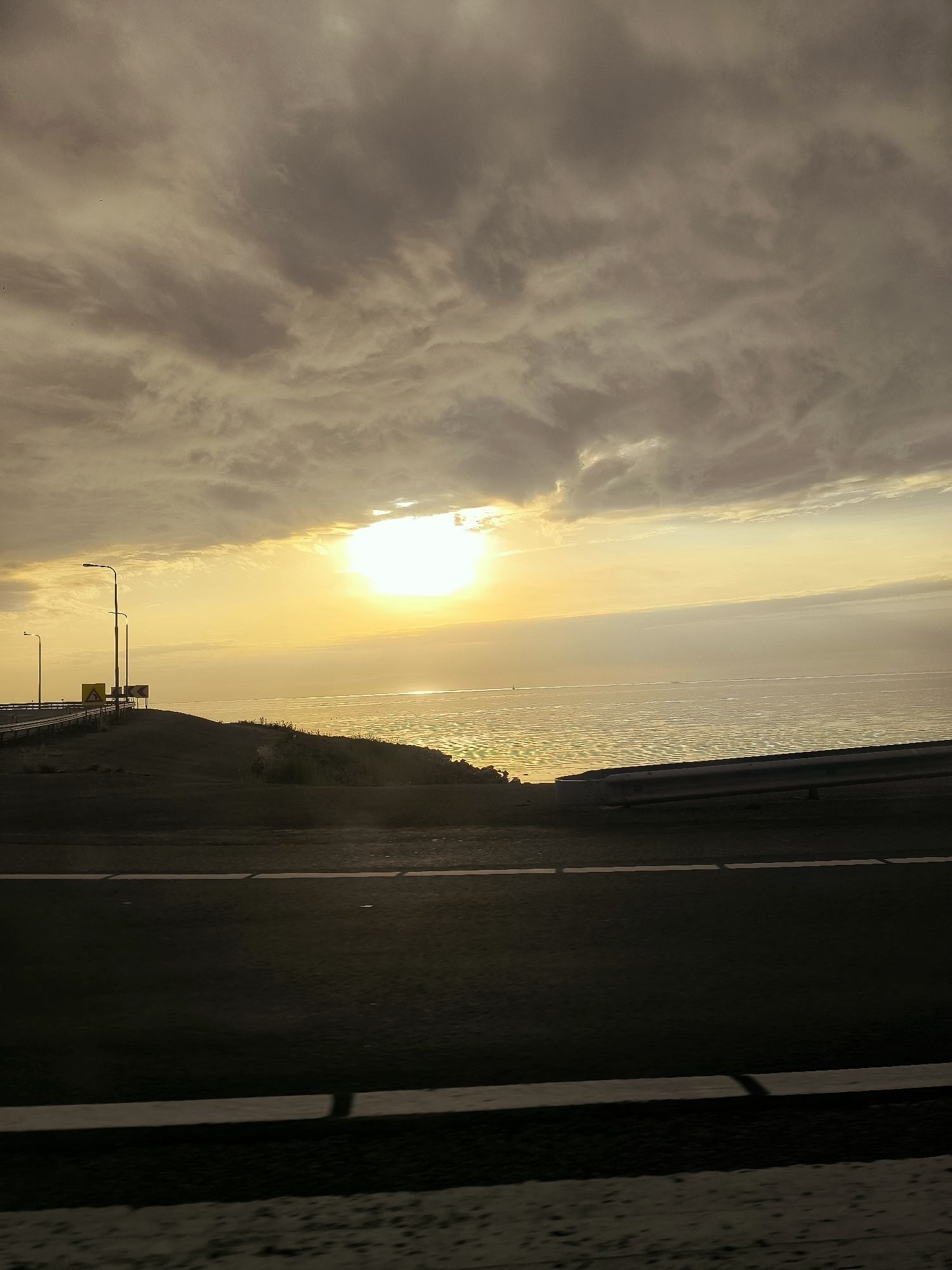 The image captures a dramatic sunset over the IJsselmeer, a vast freshwater lake in the Netherlands, likely near Lelystad. The sky is filled with heavy, grey clouds that are backlit by the intense, glowing sun, creating a luminous orange and yellow haze. The sun itself is a bright, diffused disc near the horizon, casting a shimmering, golden reflection across the water.
In the foreground, a dark, paved road with a white dividing line stretches from left to right, suggesting a highway or main thoroughfare. To the left, a raised embankment is visible, topped with sparse vegetation and a few tall lampposts. Two yellow directional signs are present on the embankment, one indicating a left curve and another a double curve to the right. A metal guardrail runs along the edge of the road on the right side. The water's edge is lined with low, dry grasses and reeds. The overall atmosphere is one of subdued natural beauty, with the harsh, overcast sky contrasting with the warm glow of the setting sun on the water. There are no people or animals visible in the scene.