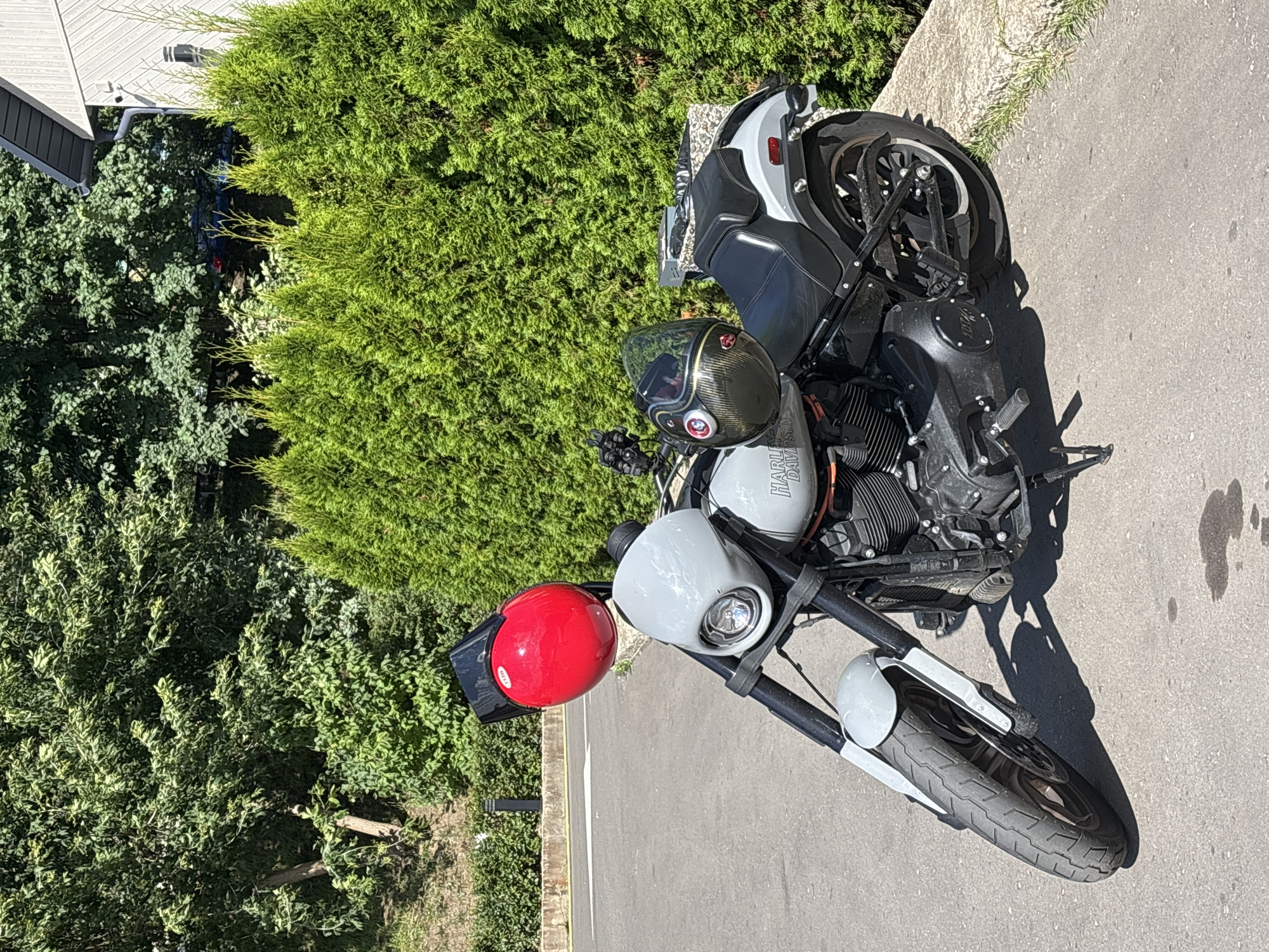 A motorcycle is parked on an asphalt surface outdoors, likely in a residential area given the presence of a house and manicured hedges. The motorcycle is a Harley-Davidson, painted a light gray color, with black handlebars and engine components. Two helmets are attached to the motorcycle: a bright red helmet is mounted on the handlebars, and a dark gray and carbon fiber patterned helmet is resting on the seat. The background features lush green trees and a modern-looking building. The scene is well-lit by daylight, casting shadows from the motorcycle onto the ground. There are no people visible in the image. The location is identified as Zvenigorod, Russia.
