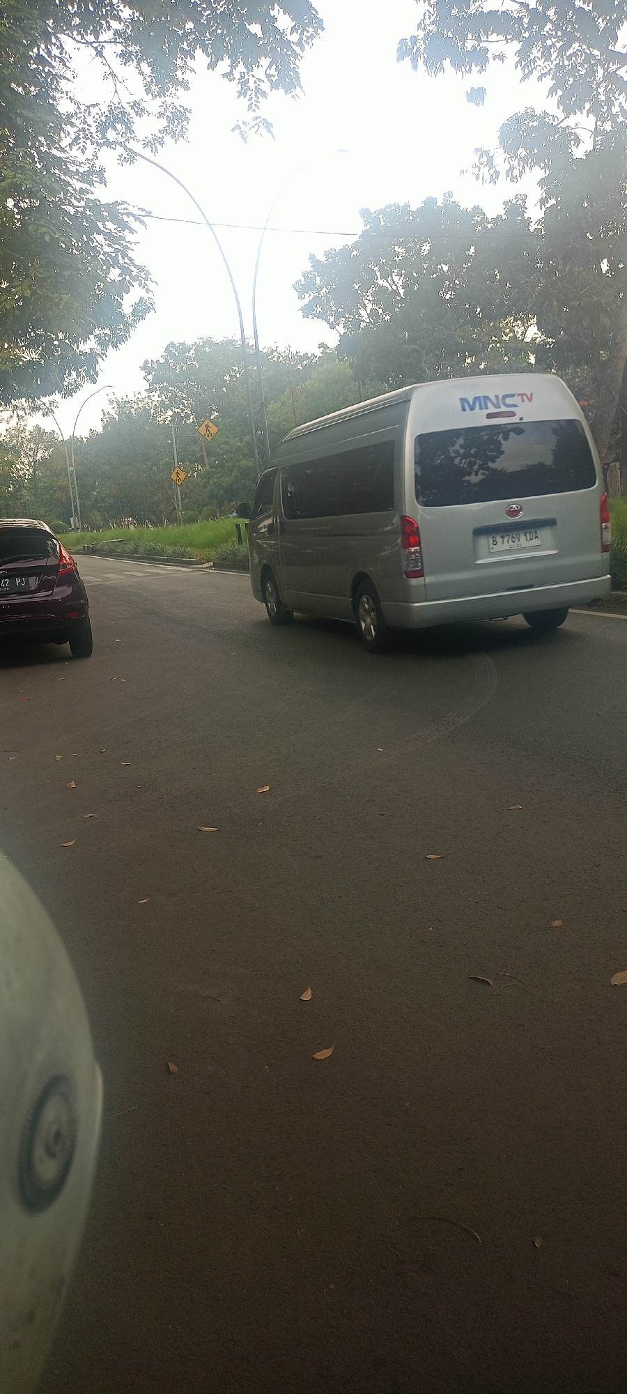 In an outdoor, asphalt road scene, a silver van with "MNC TV" branding is parked to the right, with its rear facing the camera. A dark red car is parked to the left, also with its rear facing the camera. The road has fallen leaves scattered across it. In the background, several yellow pedestrian crossing signs are visible, along with trees and streetlights. The sky is bright and overcast, with sunlight filtering through the leaves of the trees. This scene is located on a road in Bandung City, Indonesia, indicated by the license plate of the van which reads "B 7769 TDA".