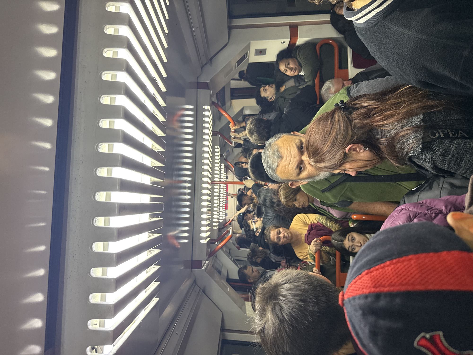 The image depicts the interior of a crowded subway car in Buenos Aires, Argentina. The scene is tightly packed with people, many standing due to the lack of seating. Several passengers are visible, including a woman with long brown hair, a man with gray hair and a green shirt, and a person wearing a black baseball cap with red accents. The subway car's lighting system is visible along the upper left side of the frame, showing a series of horizontal light fixtures. Passengers are holding onto overhead rails for support. The overall environment suggests a typical rush-hour commute.