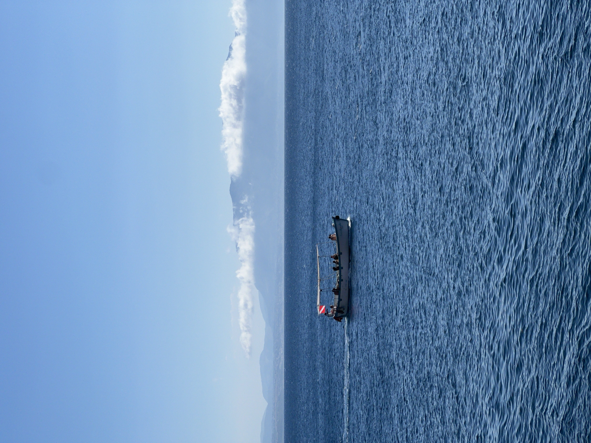 A small white boat with a canopy is traveling from left to right across a deep blue, slightly choppy sea. The boat, which has several people on board, is flying a red and white flag. In the background, a hazy, mountainous coastline is visible under a clear blue sky, with some scattered white clouds hugging the tops of the mountains. The scene suggests a sunny day at sea.
