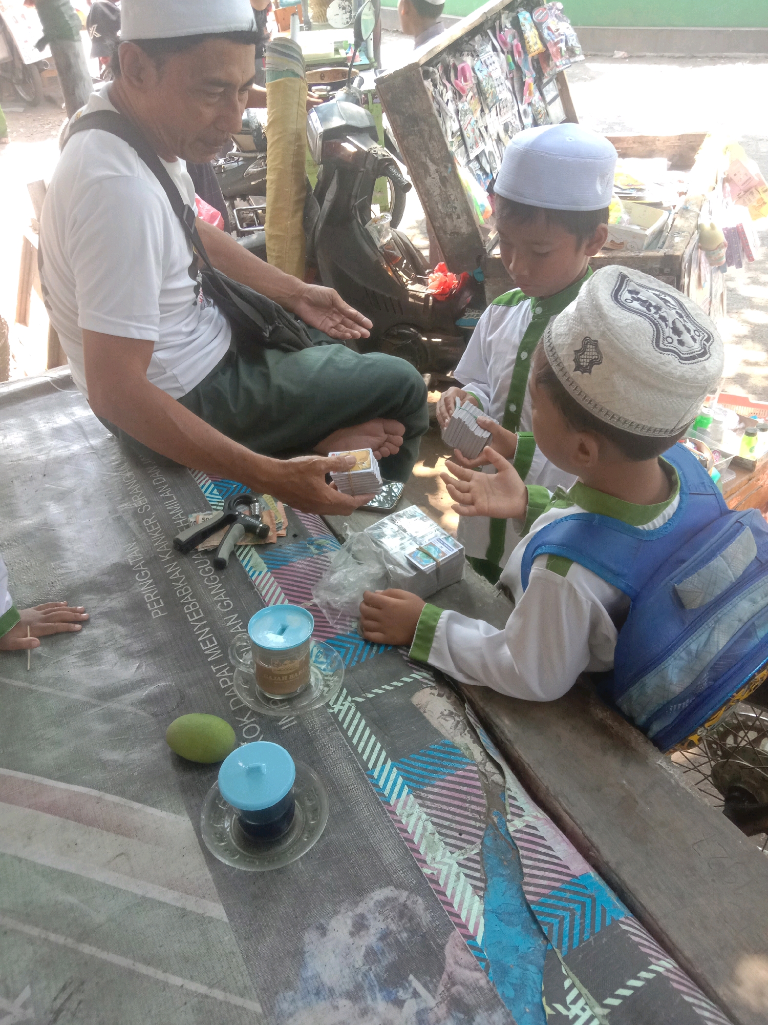 In Ngempit, Indonesia, an outdoor scene unfolds with a man and two young boys seated at a low table. The man, wearing a white t-shirt and green pants, holds a stack of small, rectangular items. One boy, dressed in a white and green outfit and a white cap, looks down at his hands. Another boy, also in similar attire and wearing a white skullcap with intricate patterns, has a blue backpack slung over his shoulders. On the table, two clear cups with blue lids sit next to a green fruit. A handwritten sign with text in Indonesian is visible beneath the table. The background shows a makeshift stall with various items displayed, and the general environment suggests a bustling, outdoor market or gathering place.