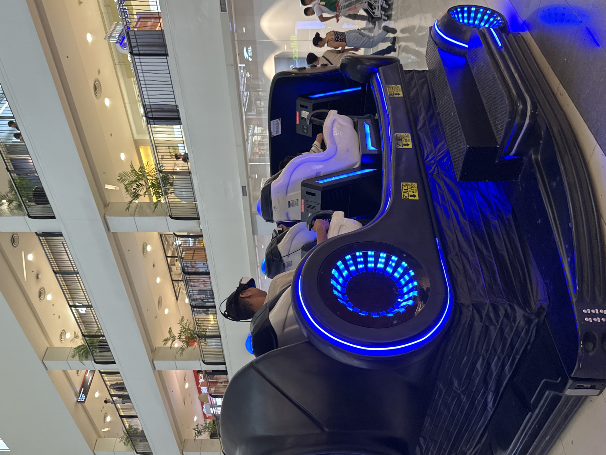 In the foreground, a black, futuristic-looking motion simulator ride is set up in what appears to be a shopping mall. The ride has two seats, one occupied by a person wearing a VR headset. The seats are white and contoured, with blue LED lights accentuating the ride's structure, particularly around the wheels and in circular patterns. Yellow caution signs are visible on the side of the ride. In the background, the shopping mall environment is visible with multiple levels, escalators, railings, and shops. Several people are walking in the background, some pushing shopping carts. The mall appears to be a modern, indoor commercial space. The overall impression is a scene within a bustling commercial hub, featuring entertainment and retail activities. This scene is likely located within a mall in Davao City, Philippines, given the context provided by the prompt.