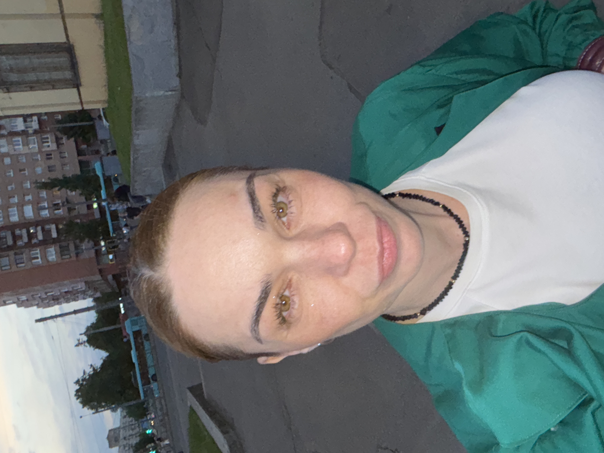 A female individual is depicted in a self-portrait style photograph, captured outdoors in an urban setting during dusk or twilight in Sinyavino, Russia. The subject occupies the majority of the frame, positioned centrally and facing directly towards the camera. She has light brown hair, parted centrally and pulled back, revealing a fair complexion with visible freckling across her nose and cheeks. Her irises are amber-colored, reflecting ambient light or small glints. Her lips are slightly pursed. She is wearing a white crew-neck t-shirt underneath an unbuttoned emerald green long-sleeved garment. A dark, possibly beaded, choker-style necklace with small gold-colored accents is visible around her neck.
The background depicts a city street scene. To the left of the subject, a light blue public transit bus is visible on an asphalt roadway, moving away from the viewer. Several indistinct pedestrian figures are present on the sidewalks. Further back, multi-story residential buildings, primarily constructed of reddish-brown brick, are visible. Utility poles and overhead wires are present against a cloudy sky. To the right of the subject, a two-story building with a light yellow facade features dark, possibly boarded-up or covered window openings on its upper level, above a patch of green grass on a raised concrete curb. The street surface shows some cracking. The ambient light indicates low light conditions, consistent with evening.