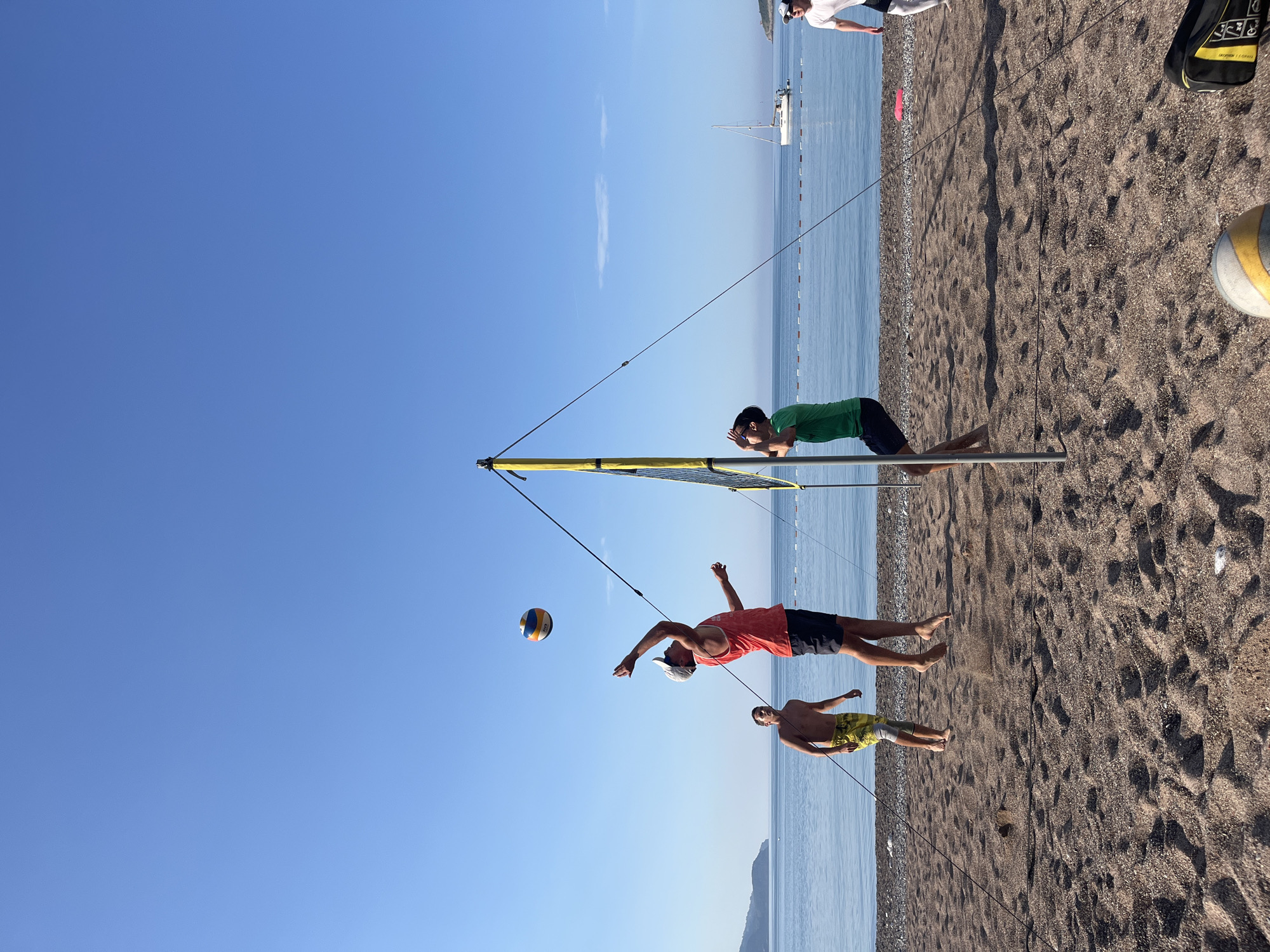 The image captures a beach volleyball game in Bečići, Montenegro. Two teams of two are playing, and a player in an orange shirt, black shorts, and a white cap is jumping in the air to hit the volleyball. Another player in yellow shorts is nearby. Across the net, a player in a green shirt and navy shorts leans on the net with his hands on his face, seemingly observing. The volleyball net is set up on the sandy beach, and the Adriatic Sea is visible in the background. The sky is clear and blue. In the distance is a sailboat and some beachgoers. The ground is made of sand. A volleyball is on the sand at the bottom right corner of the image.