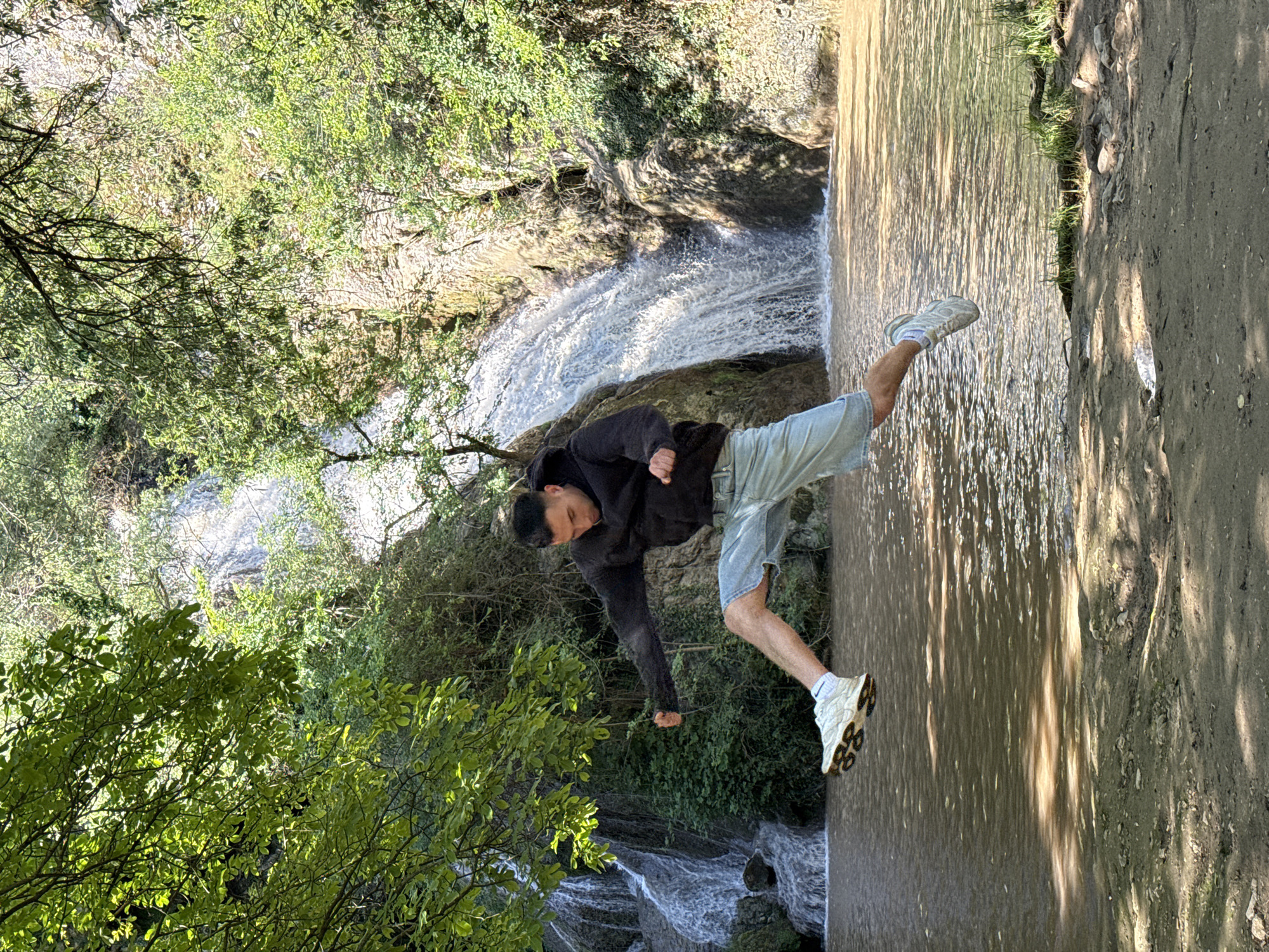 A man in a black jacket and blue shorts is standing in the water near a waterfall in Hotnitsa, Bulgaria. He is balancing on one leg with his right leg extended and is making a fist with his left hand. The area is surrounded by trees and greenery. A dirt path is visible next to the water.