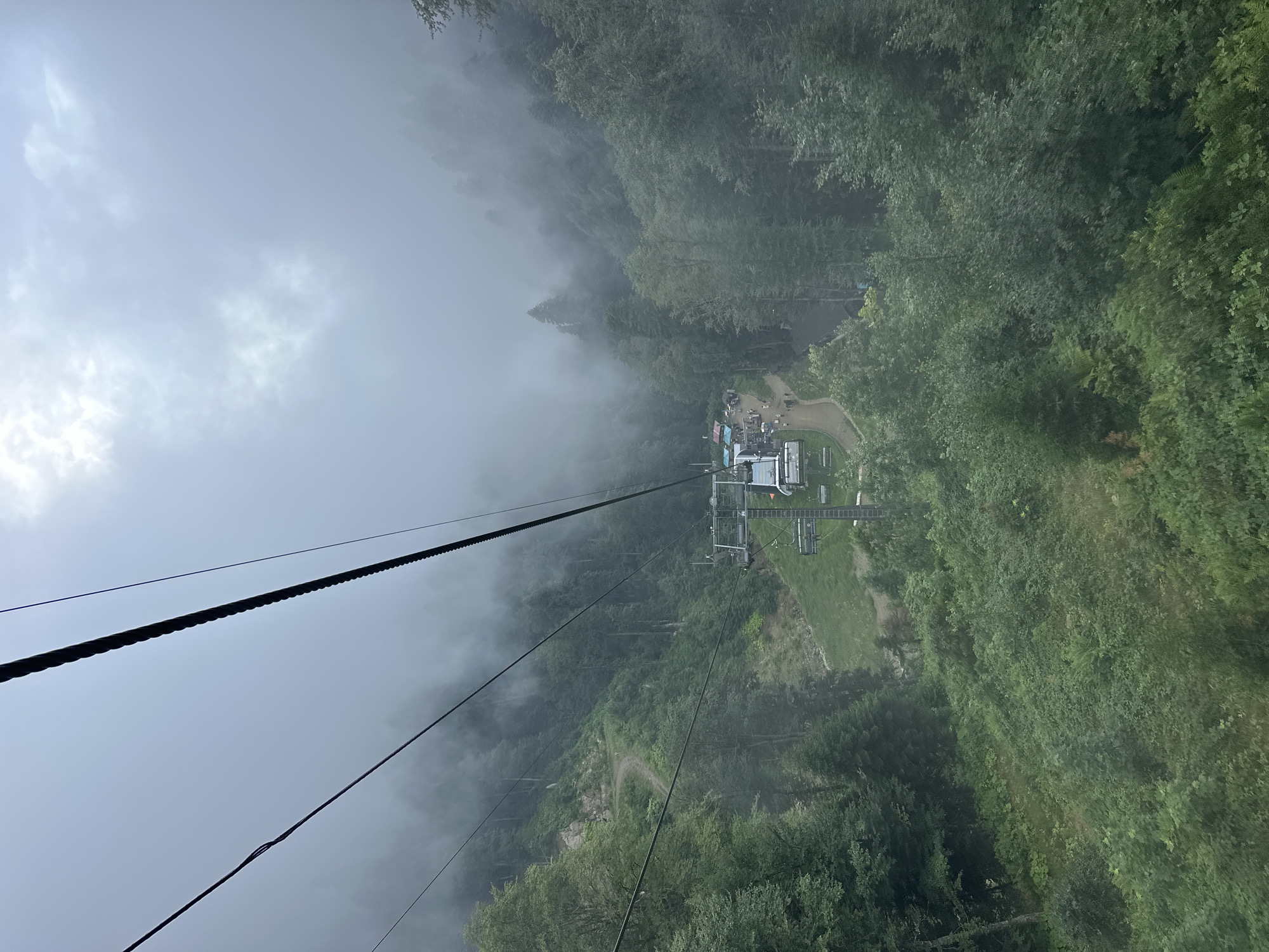 The image shows a view from a cable car descending in a mountainous, forested area. Thick fog obscures the upper portions of the trees and the sky. Below, a cable car station is visible with several people and small structures. A winding dirt path leads away from the station into the dense green foliage. The scene appears to be located in the resort area of Estosadok, Russia, likely at a higher elevation due to the cable car and mountainous terrain. The overall impression is one of a misty, natural landscape with signs of human activity at the base station.