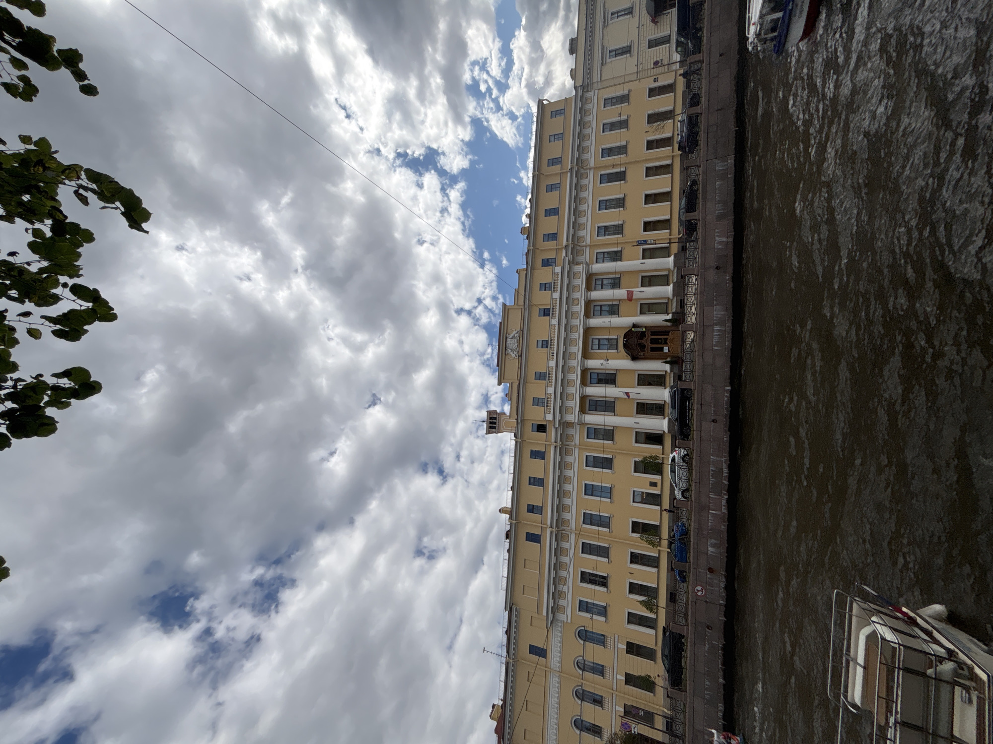 The image depicts a wide, brown river in the foreground, with a white boat in the lower left corner. The river flows in front of a large, yellow neoclassical building with a colonnade. The building has numerous windows and a central entrance with a wooden door. The sky above is dramatic, filled with a mixture of white and grey clouds, with patches of blue sky visible. Green tree leaves hang from the top edge of the frame, suggesting the photo was taken from beneath trees. Cars are parked along the embankment in front of the building. This scene is likely located on one of the many canals and rivers in Saint Petersburg, Russia, given the architectural style of the building and the presence of boats.